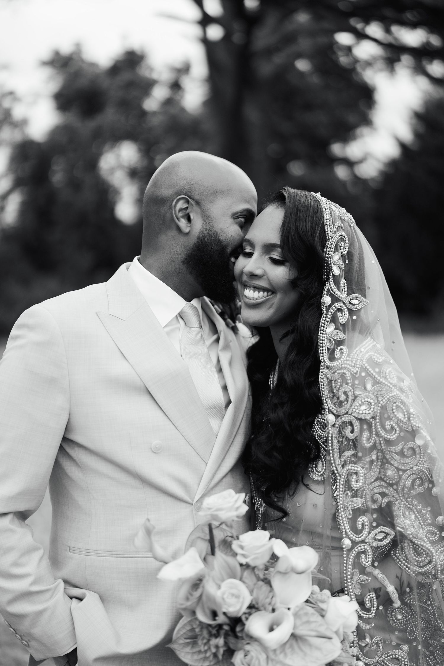 Black and white wedding photo of smiling bride in embroidered veil holding bouquet while groom whispers in her ear.