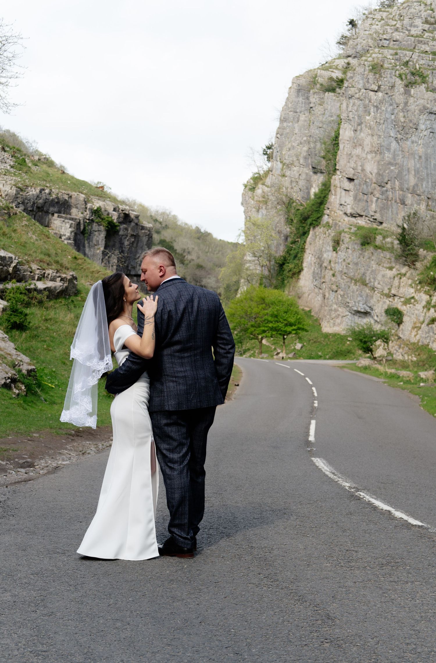 Newlyweds sharing intimate moment on mountain road with dramatic cliffs.