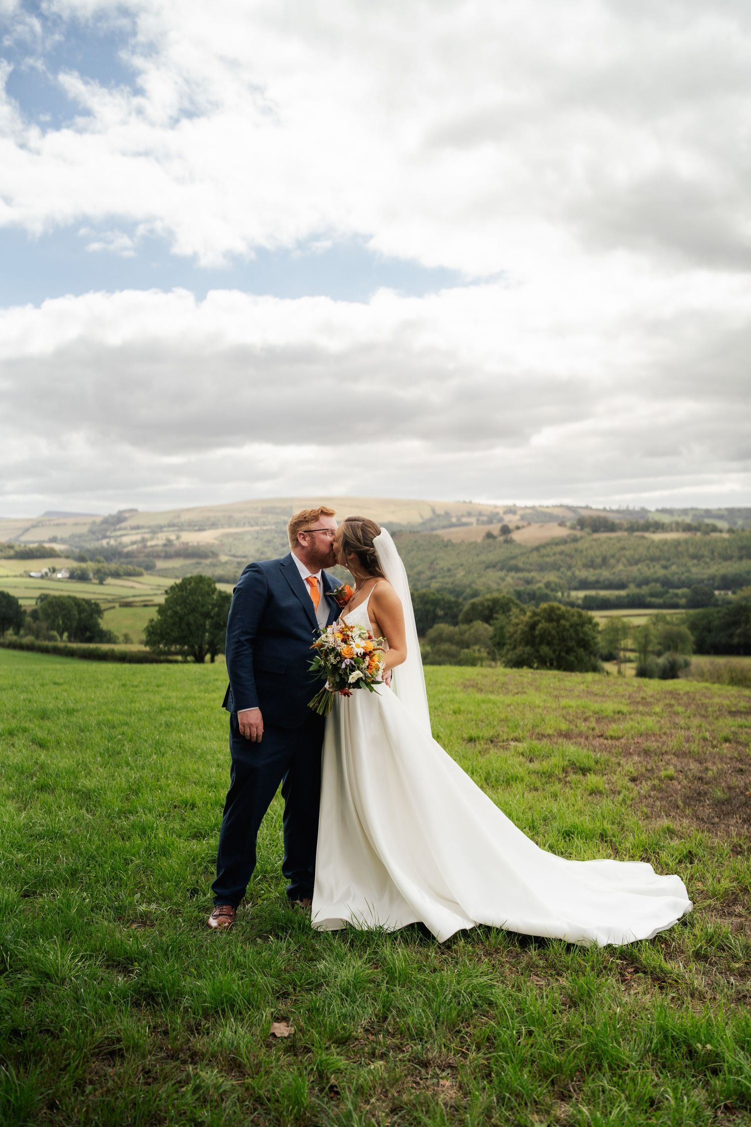 Bride and groom kissing in a green field with rolling hills and cloudy sky backdrop on their wedding day.