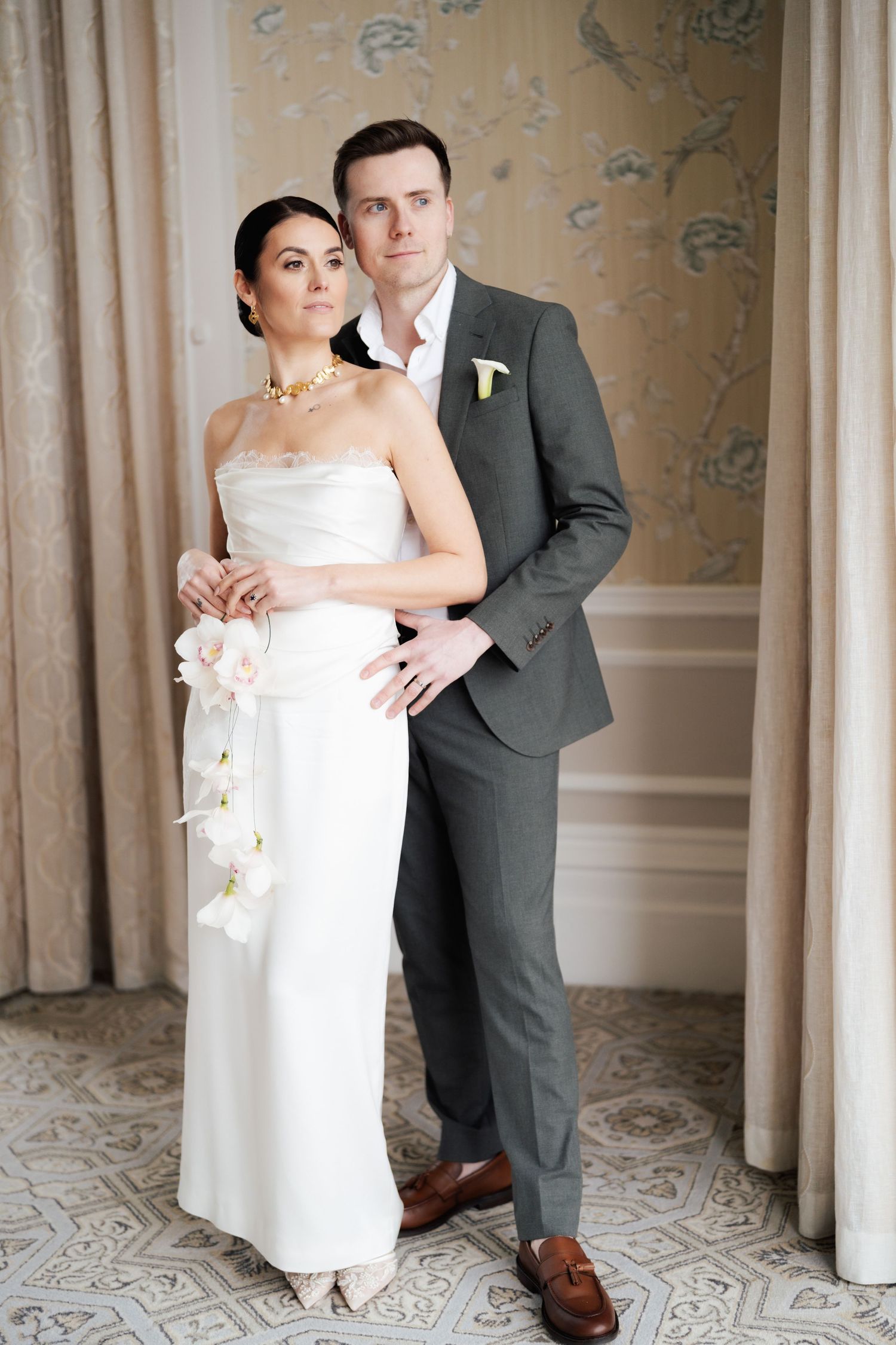 Elegant wedding couple poses together in formal attire against a wallpapered interior with ornate floor patterns.