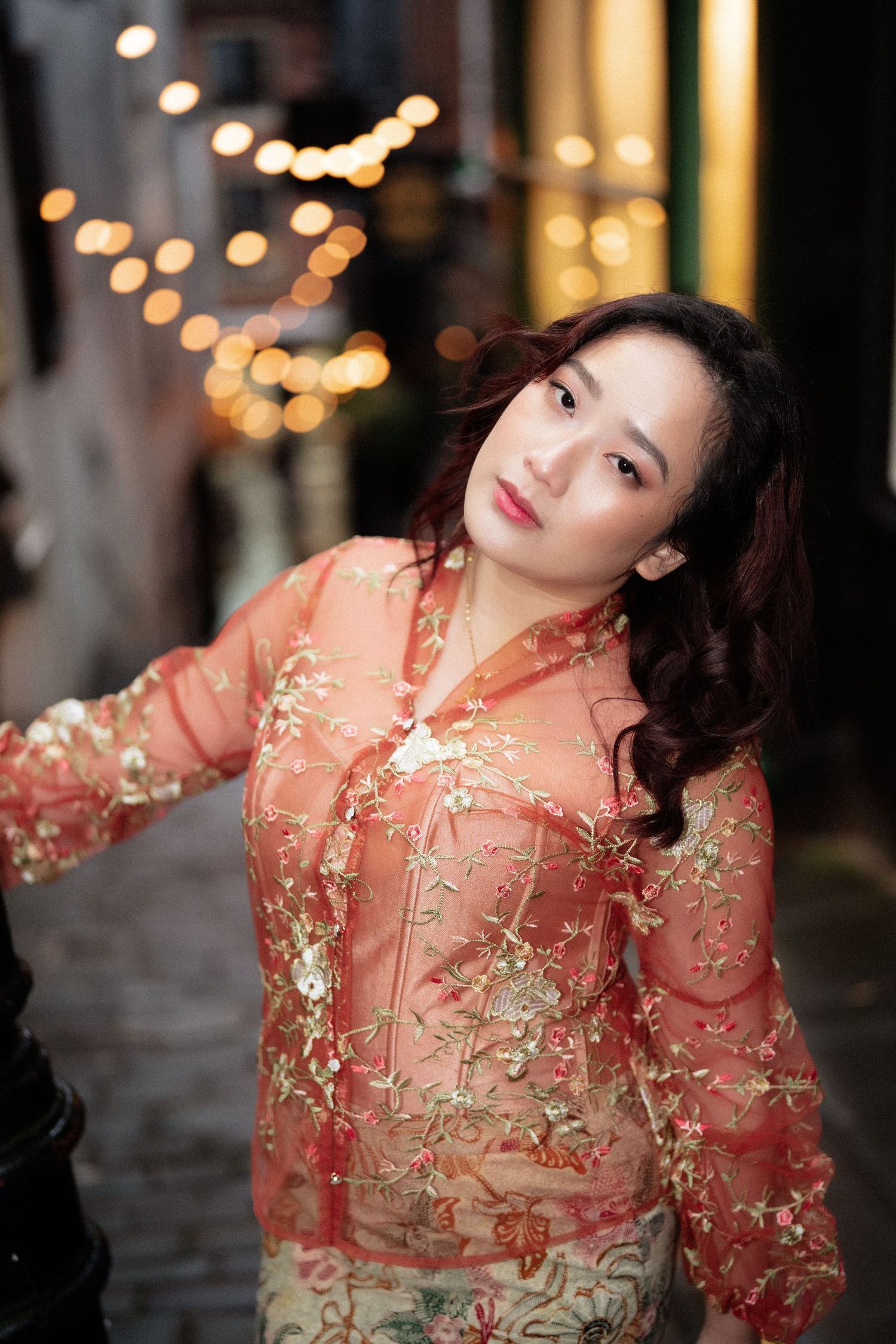 A person in a sheer coral floral dress poses gracefully against a blurred background with warm lighting.