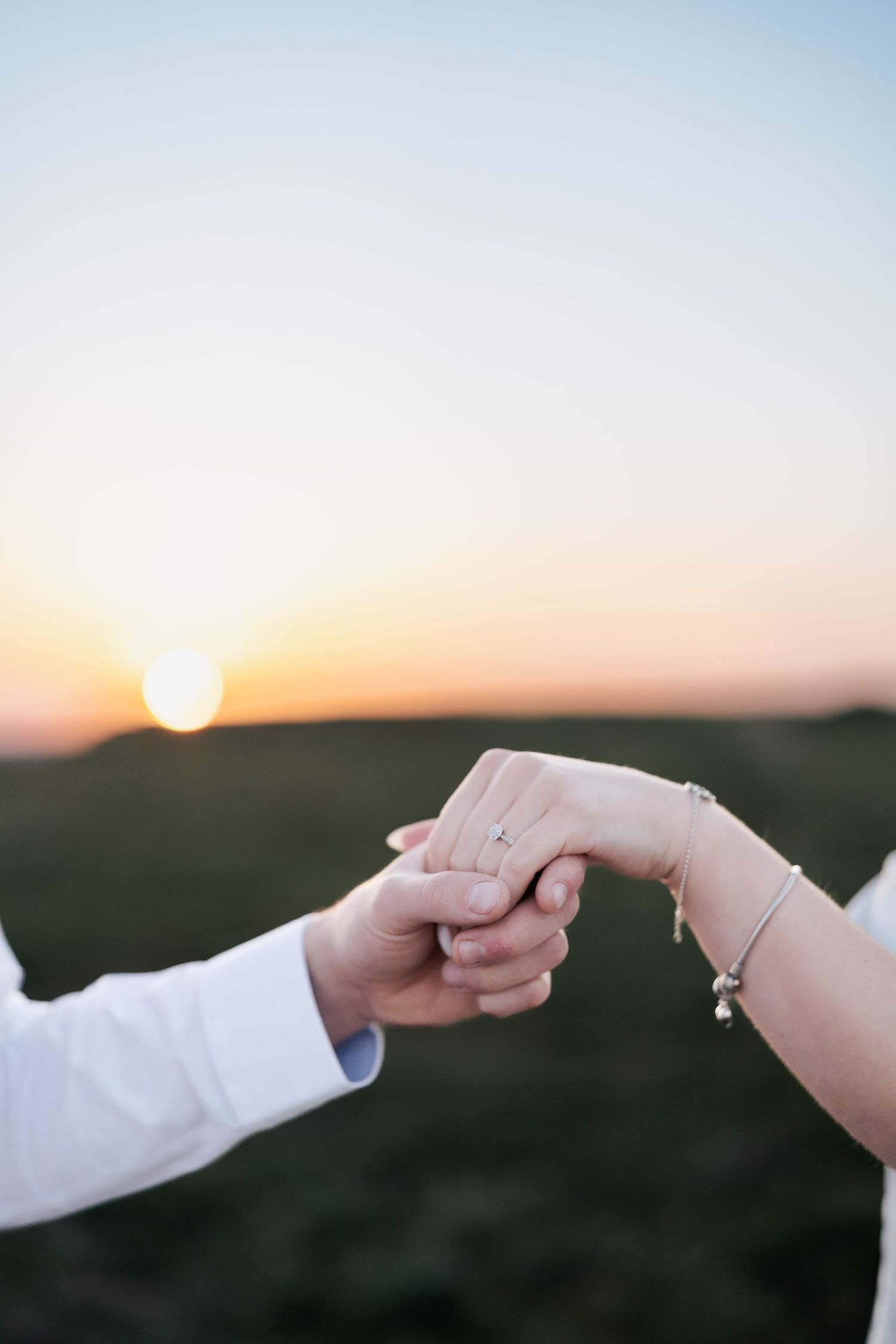 Couple holding hands tenderly at sunset in an open field, woman wearing a delicate bracelet and ring.