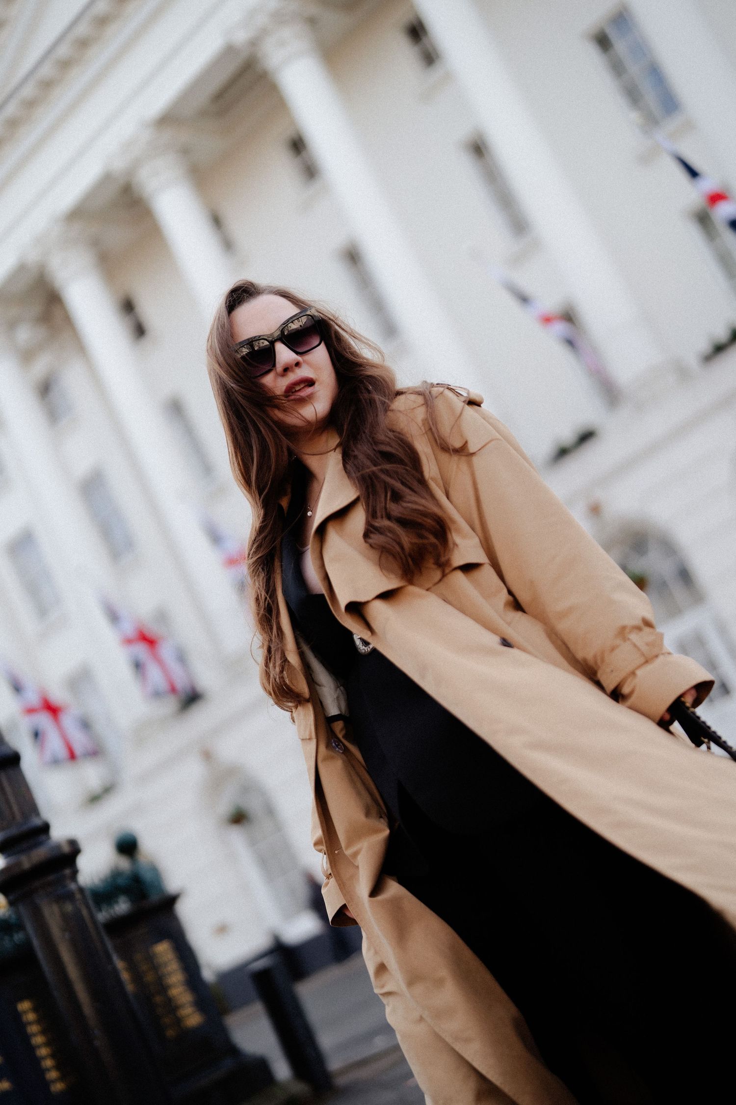 Stylish woman in beige trench coat and sunglasses posing outside a grand white building with flags.