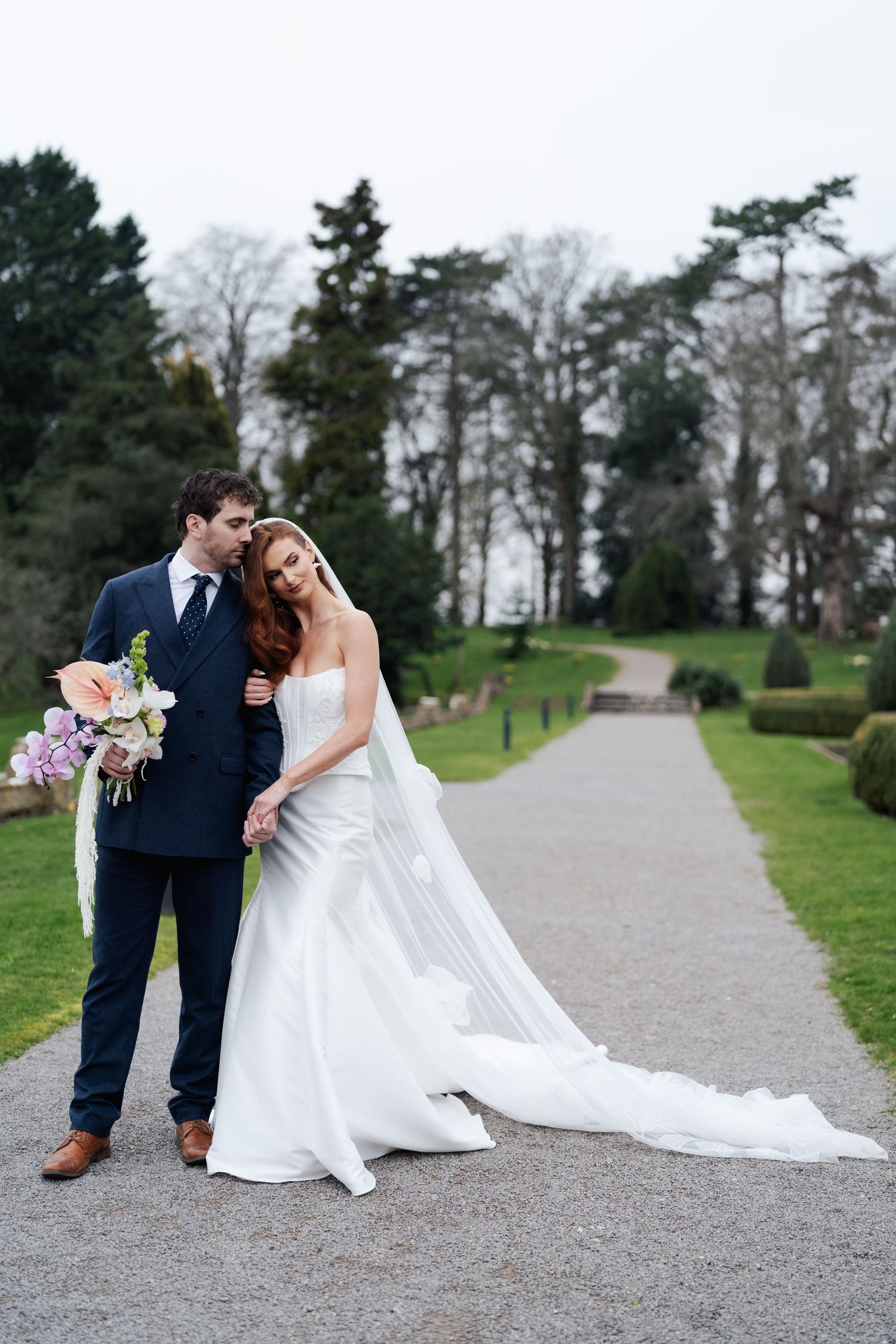 A romantic couple in wedding attire poses together on a tree-lined gravel path during their outdoor photo session.