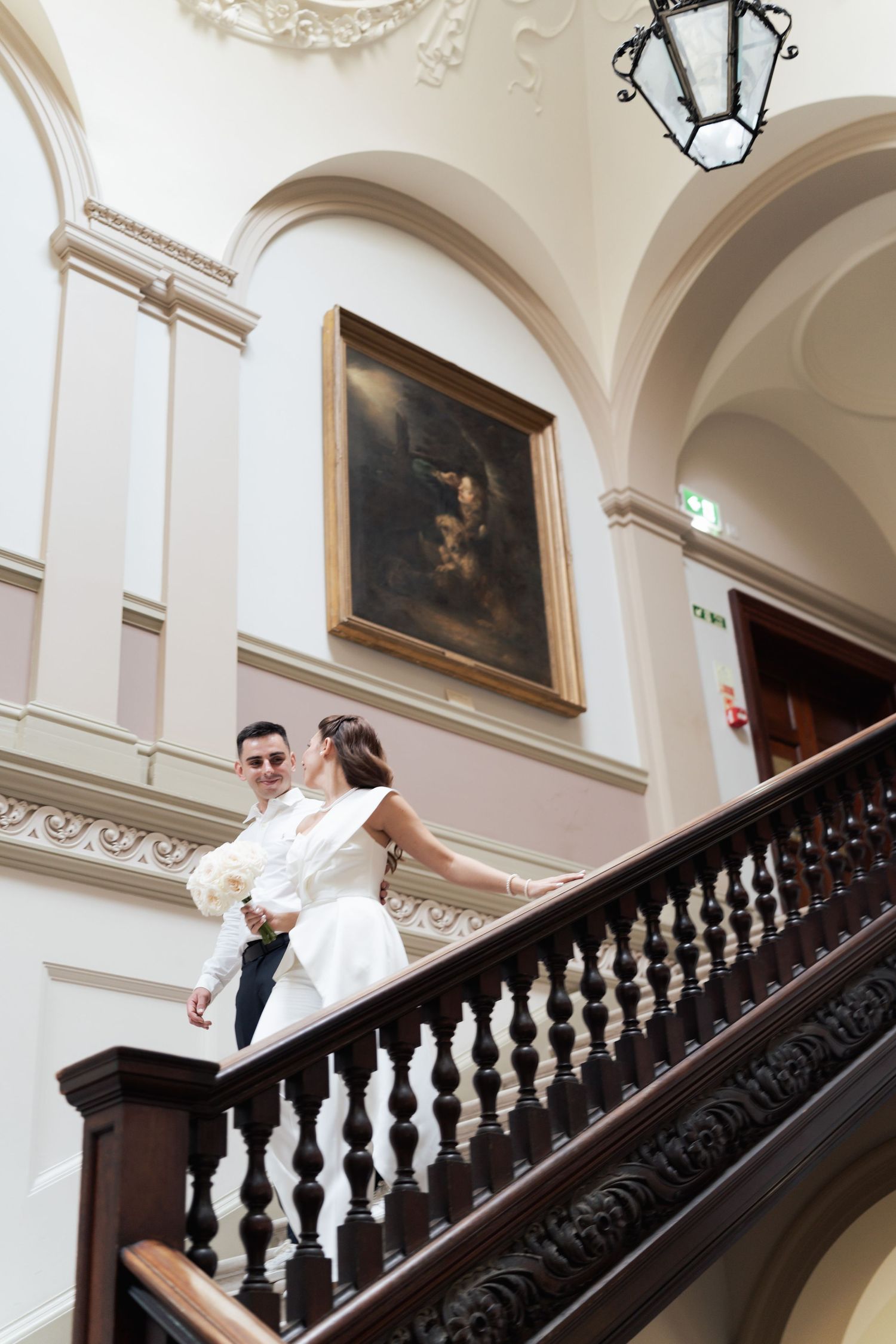 Bride and groom share a romantic kiss on an elegant staircase in a grand historic venue with arched ceilings.