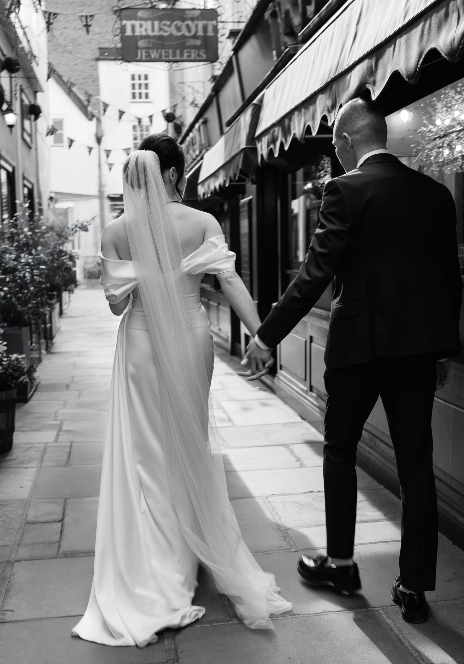 A black and white photo series showing a bride and groom walking together on a sidewalk past storefronts.