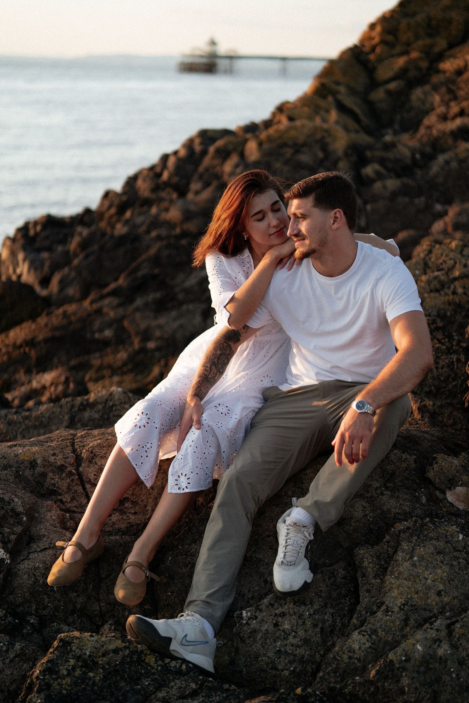 Couple in white outfits sitting intimately on coastal rocks during golden hour.