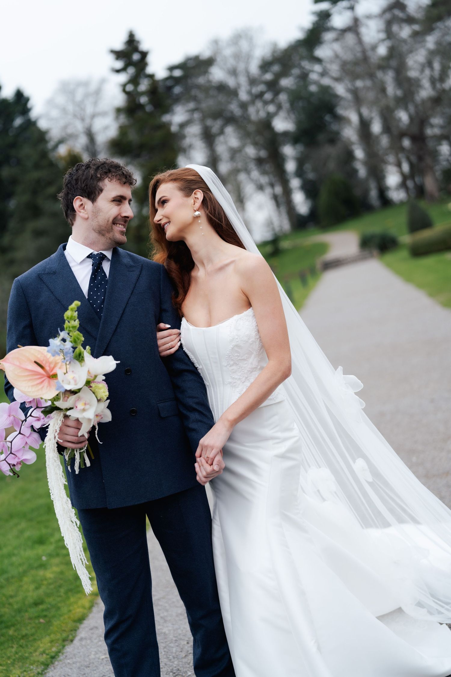 Newlywed couple in navy suit and white wedding dress share intimate moments along a tree-lined path.