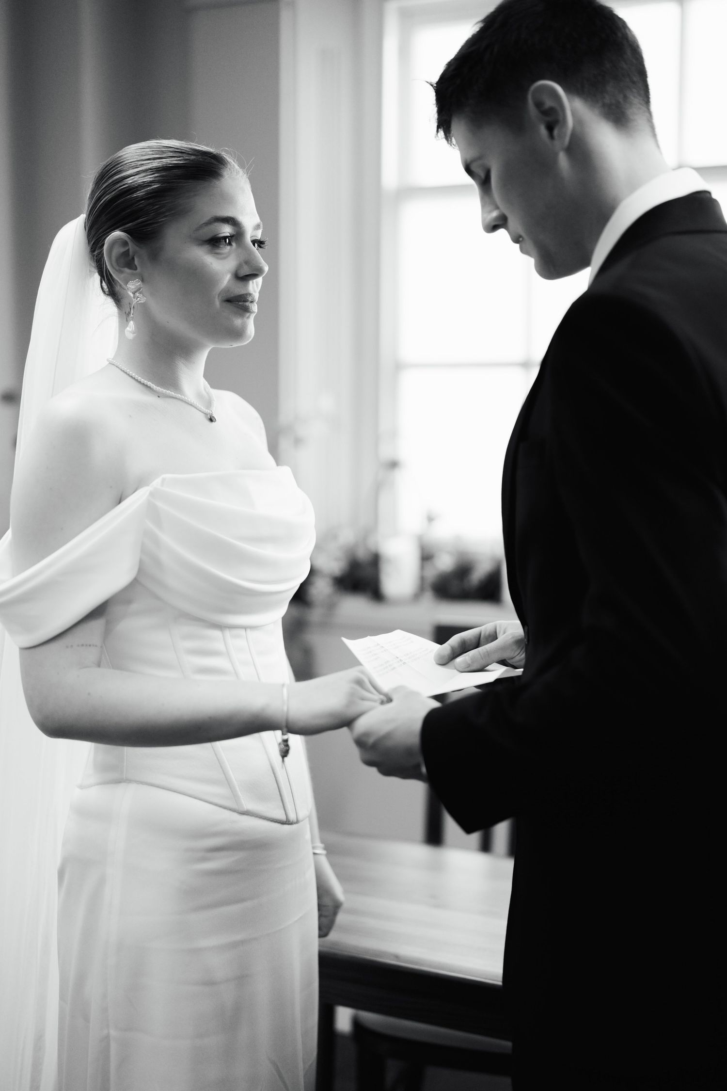 Black and white photo of bride and groom exchanging vows, bride in elegant off-shoulder gown holding written vows.