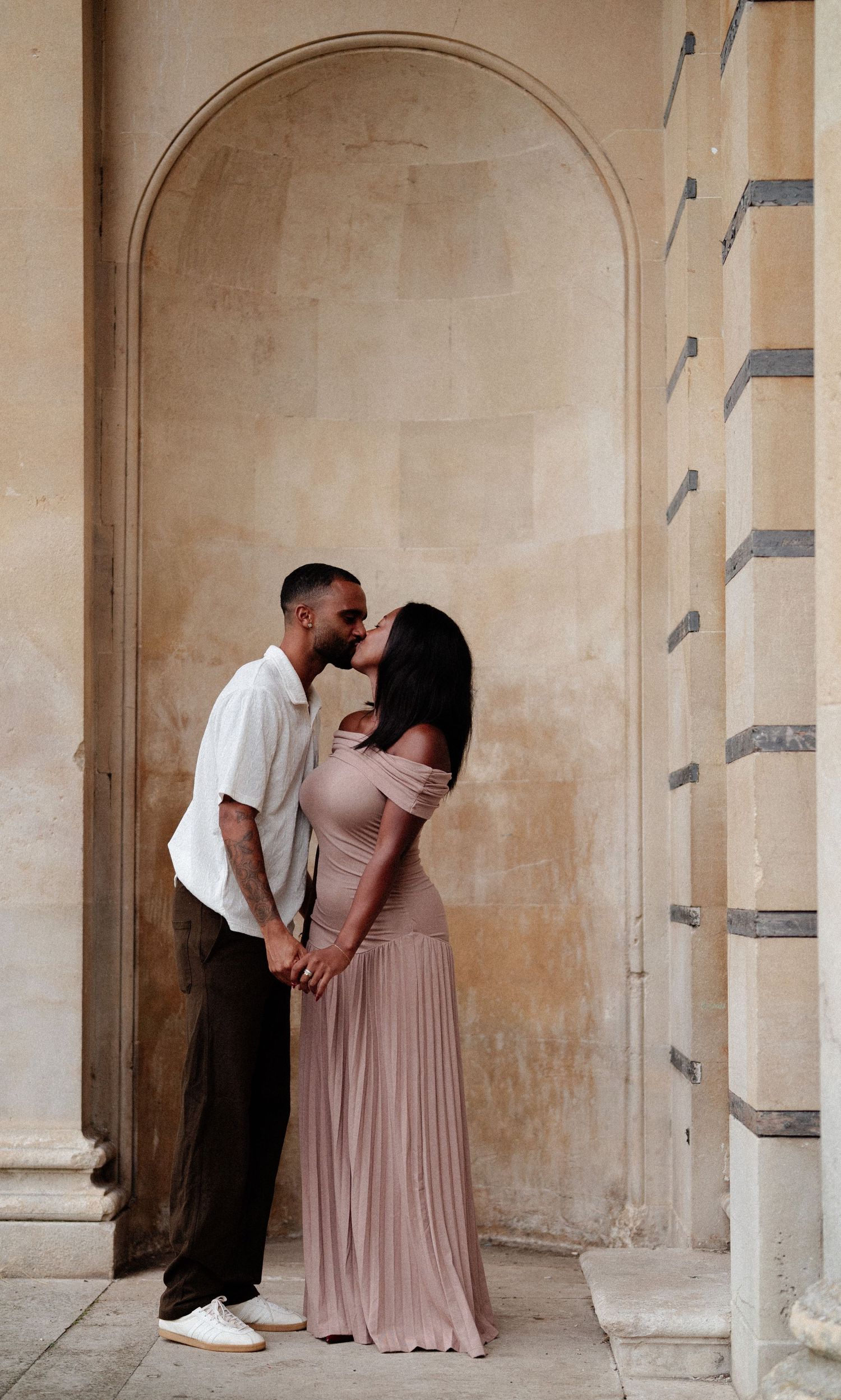 A couple shares a romantic kiss in an elegant arched alcove, she wears a blush off-shoulder gown while holding hands.