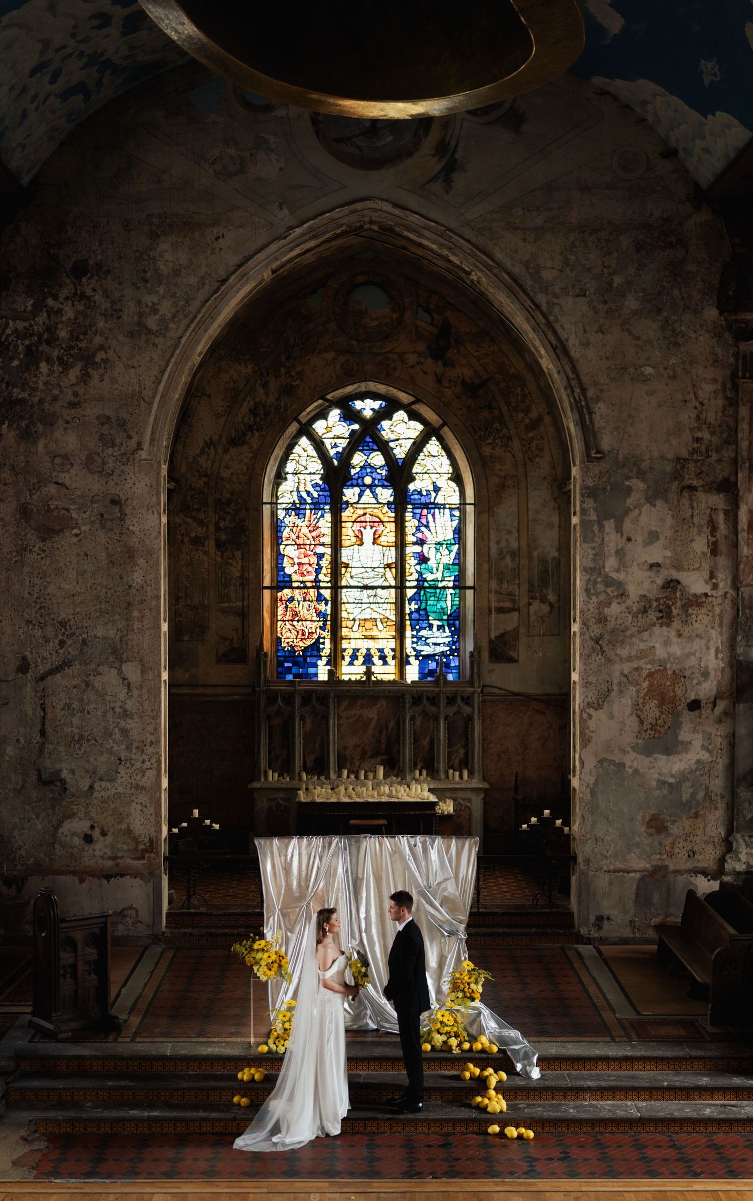 Bride and groom stand beneath a draped arch with yellow flowers before a stained glass window in a rustic church.