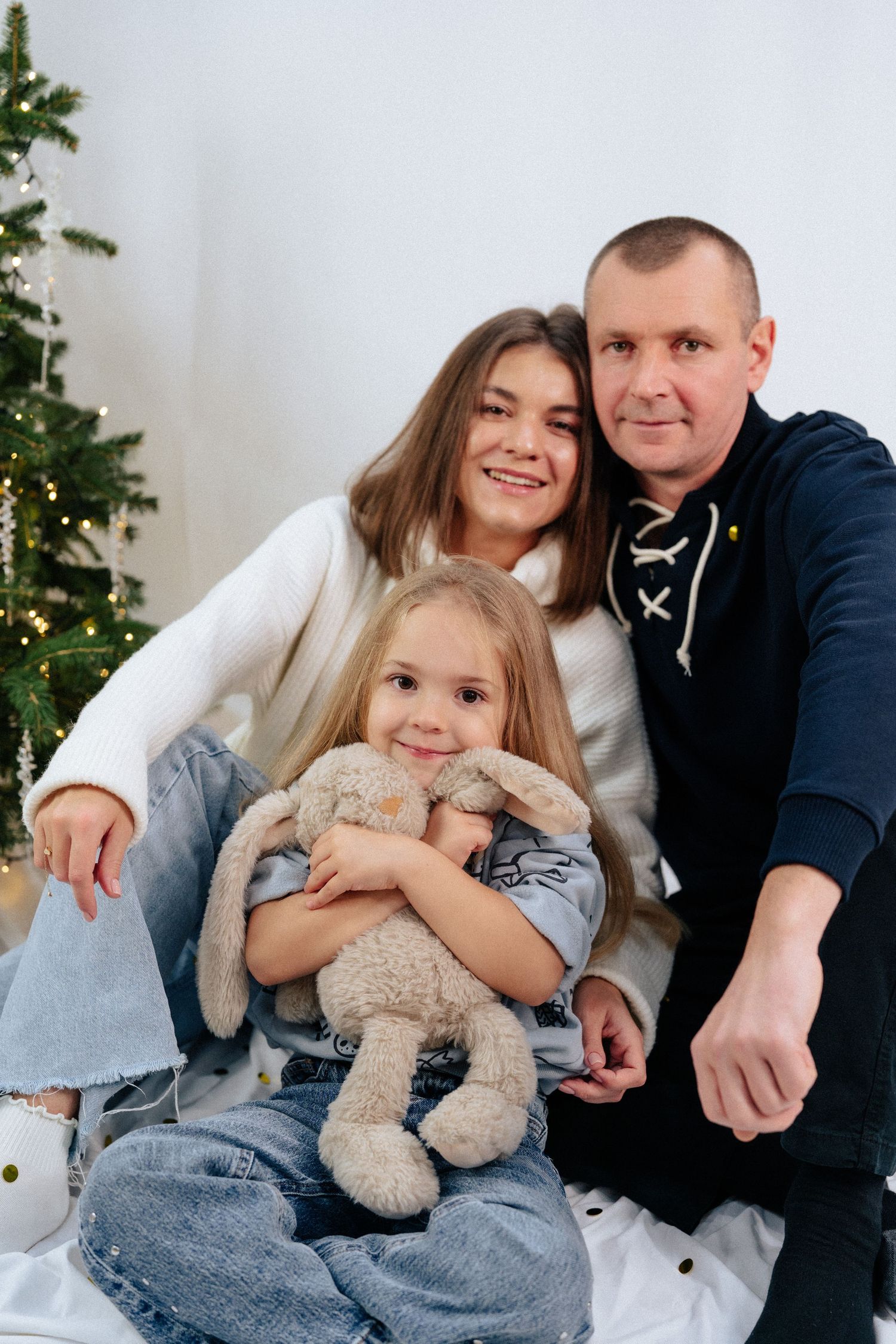 A family poses together on white backdrop with Christmas tree, holding a plush bunny toy.