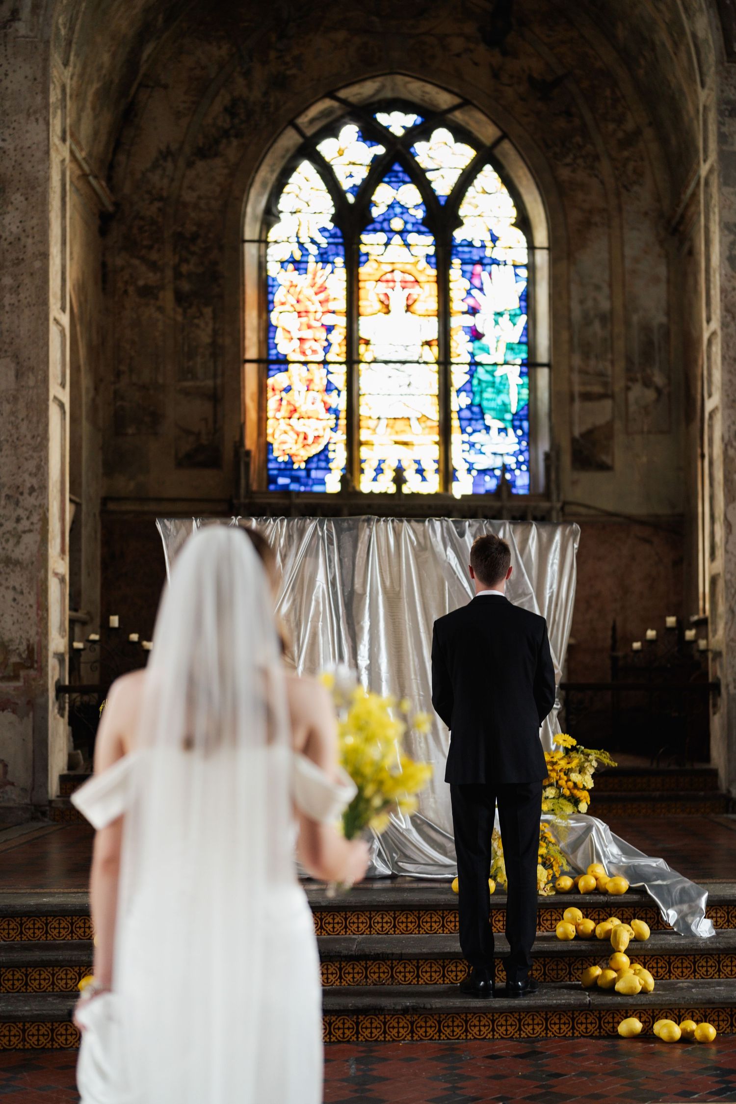 Bride and groom standing before a stunning stained glass window in a church, decorated with yellow flowers and draped fabric.