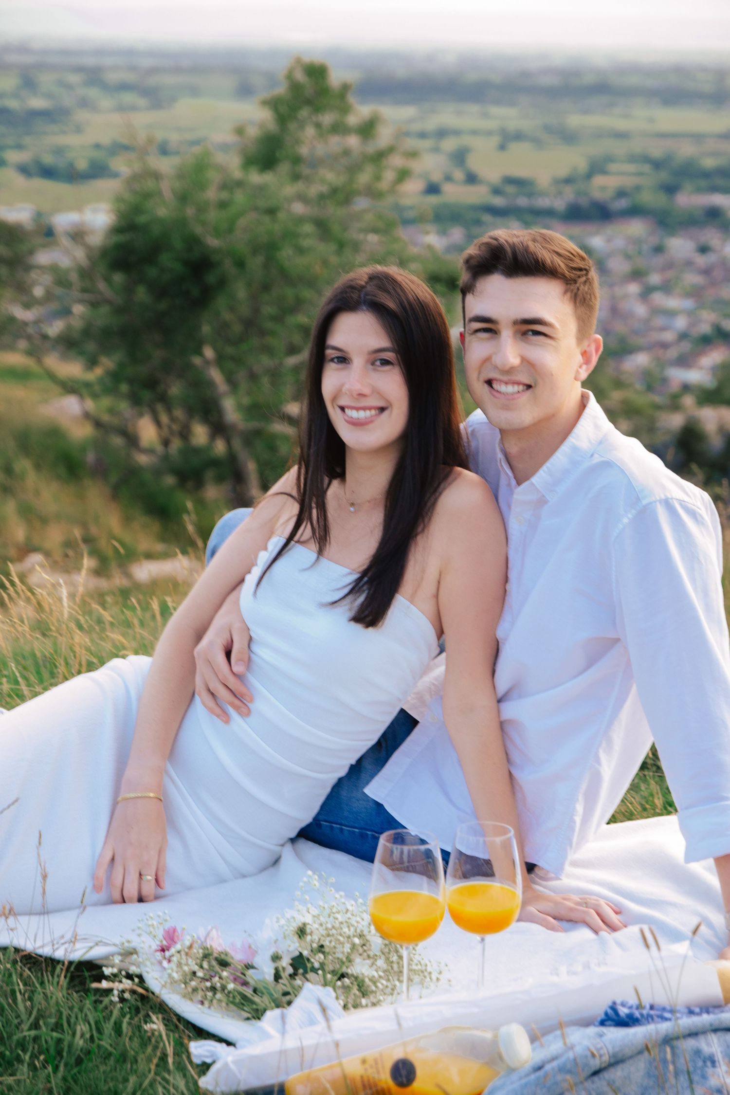 Couple in white outfits enjoying a picnic with orange juice on a hillside overlooking a town.