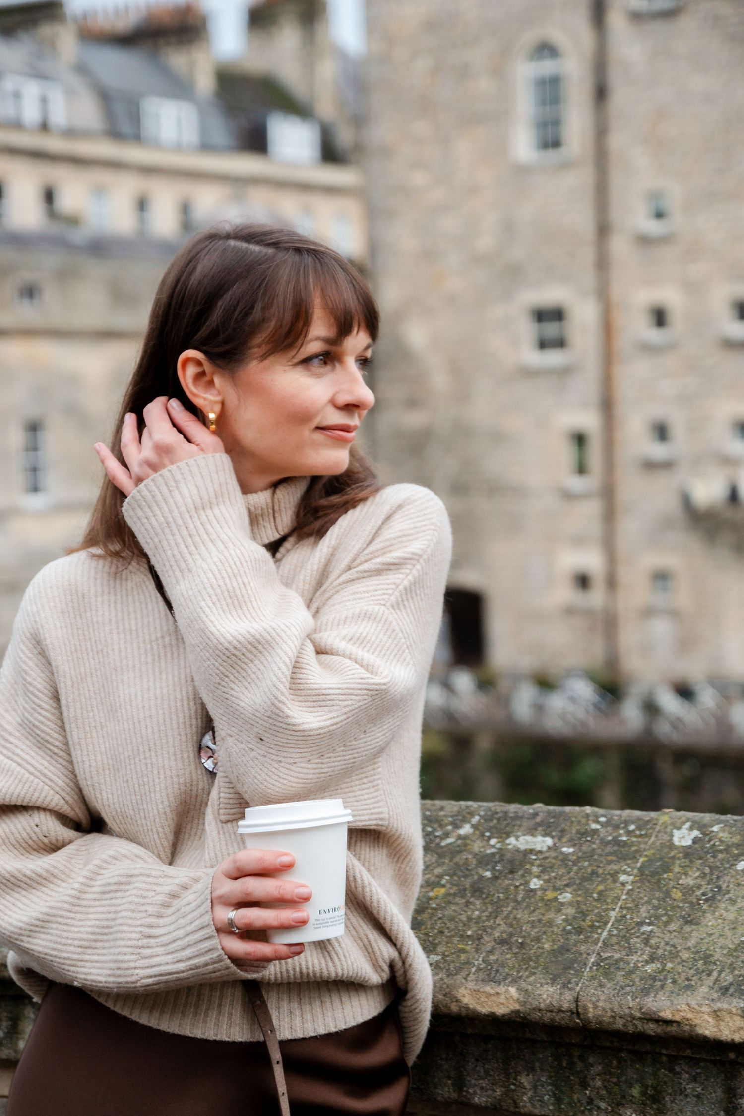 Person in cream turtleneck sweater holding cup while leaning on stone wall.