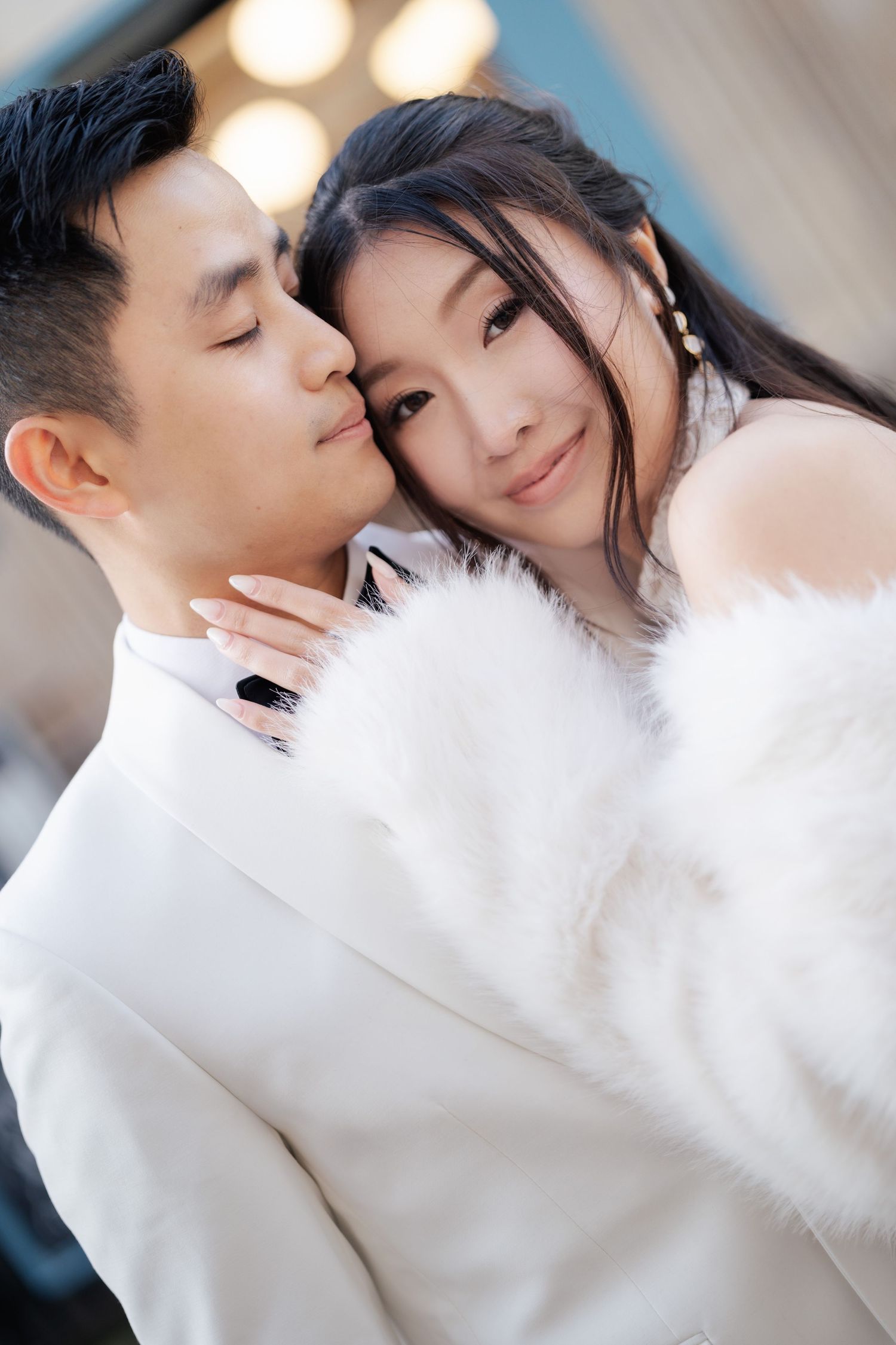 A romantic couple in white outfits embrace tenderly, with the man nuzzling the smiling woman wearing a fur stole.
