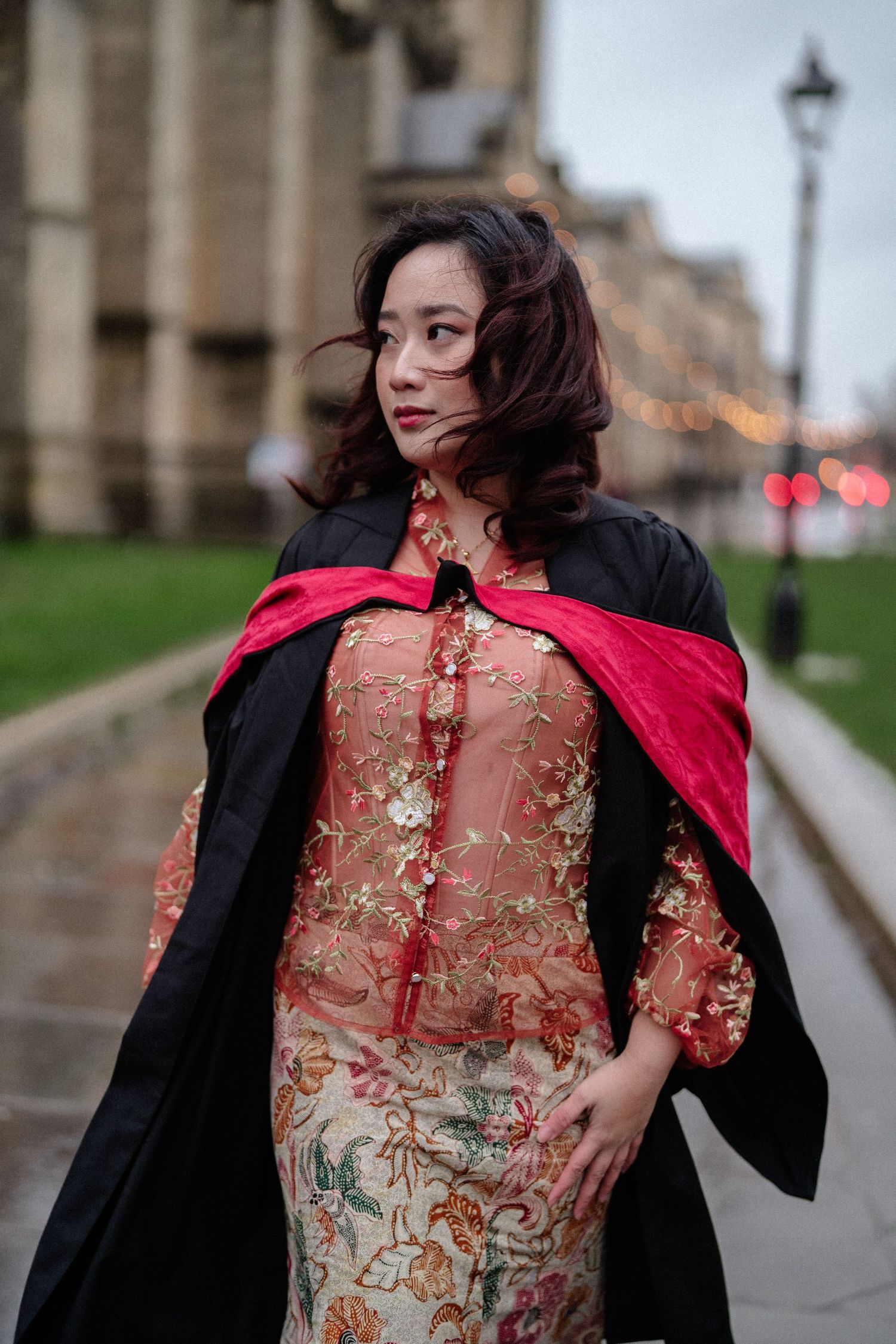 A person in a black jacket and sheer red embroidered dress poses outdoors against a blurred urban backdrop.