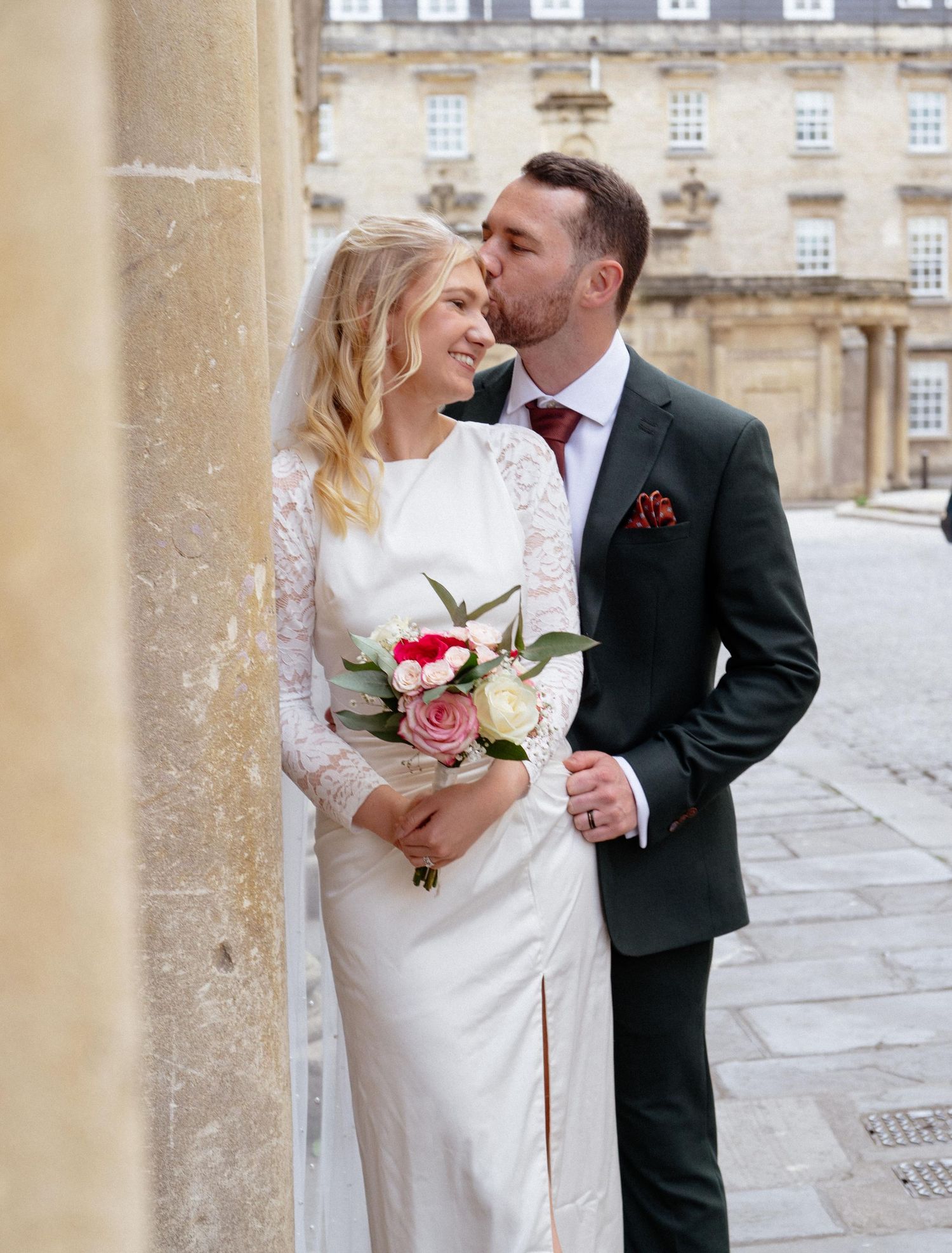 A groom kisses his bride on the cheek as she smiles, holding a pink and white bouquet, in front of a stone building.