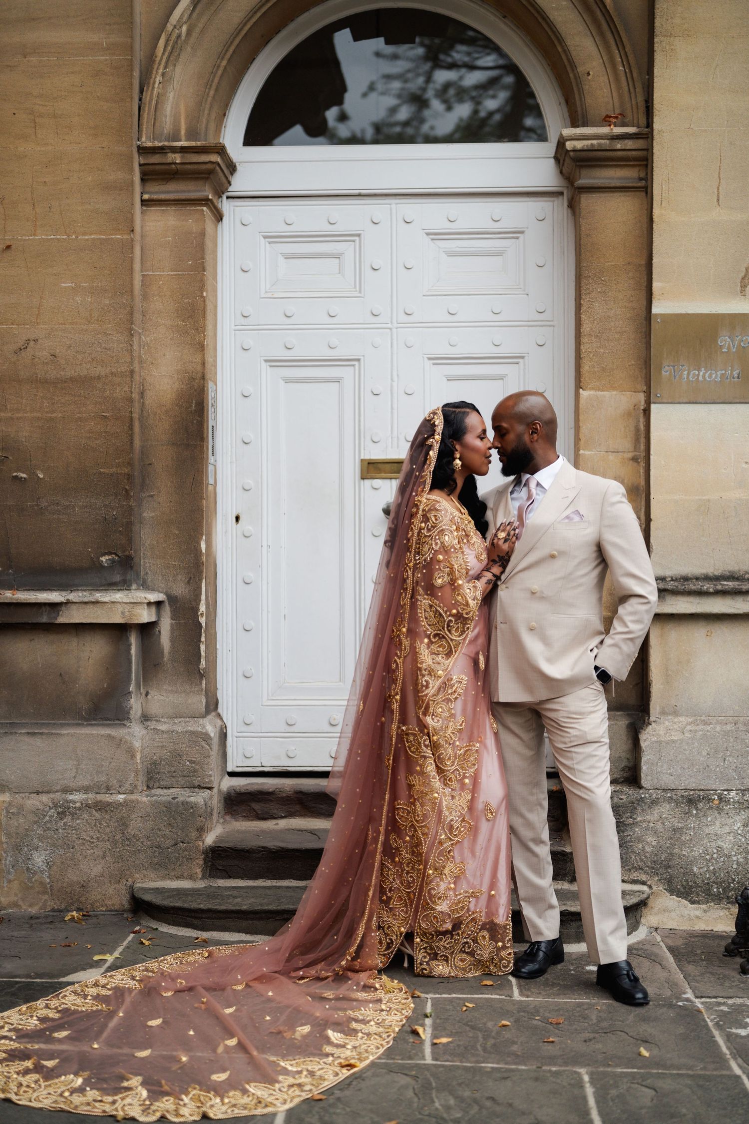 Bride in gold embroidered lehenga with pink veil and groom in beige suit share romantic moment at ornate white door.