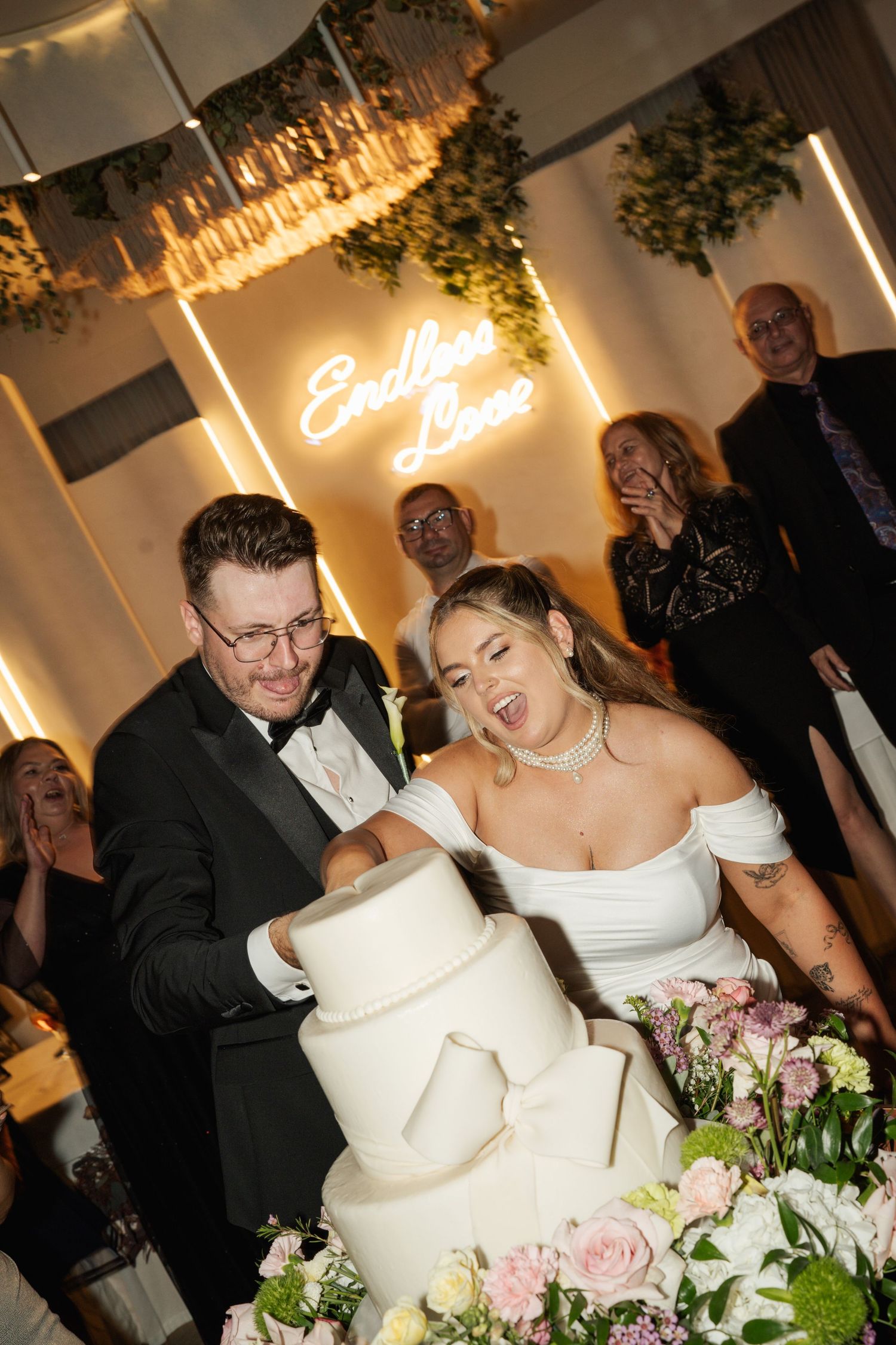 Bride and groom joyfully cutting a white tiered wedding cake with neon sign and floral arrangements.