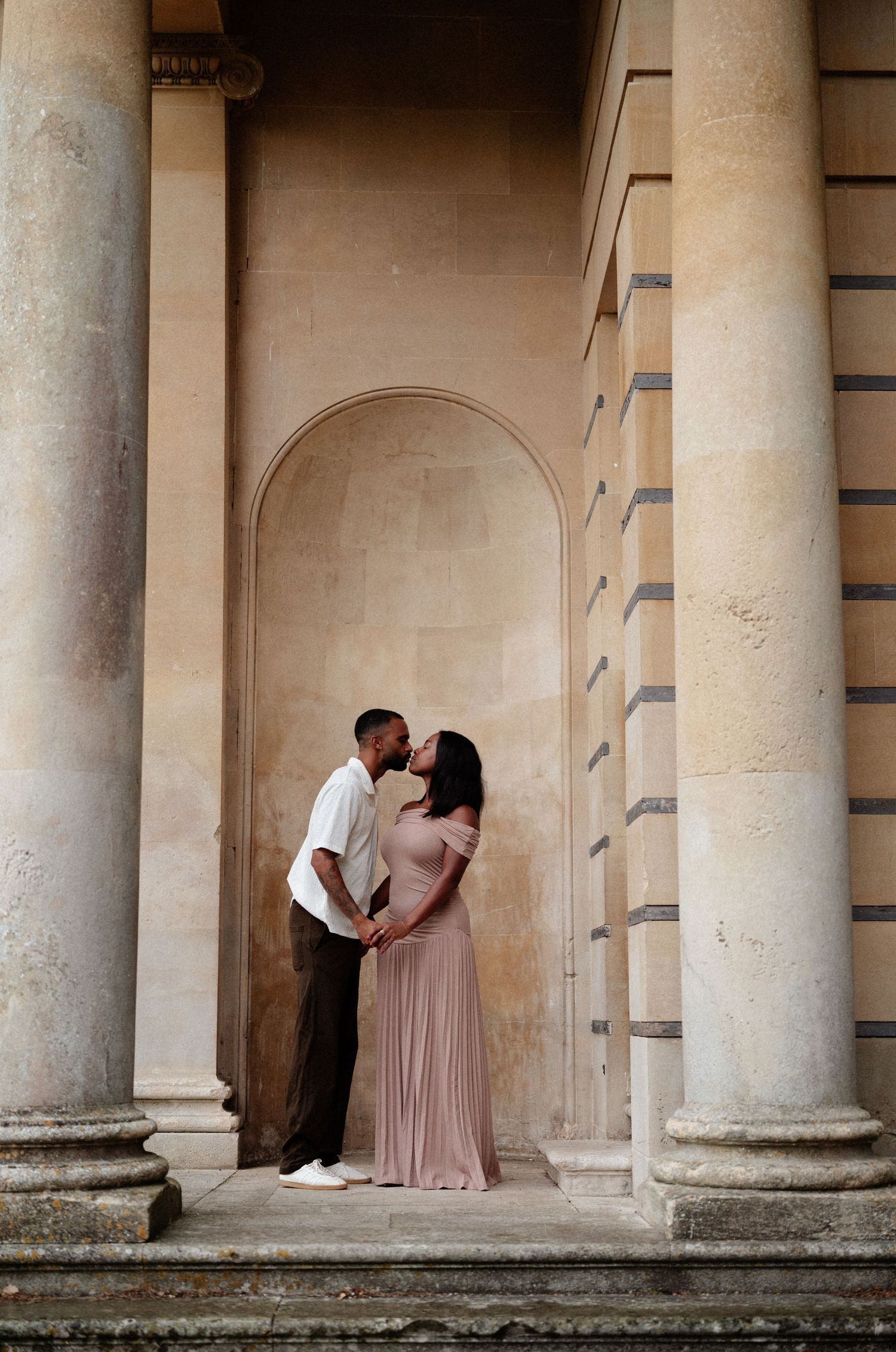 A couple shares an intimate moment between classical stone columns of a grand building.