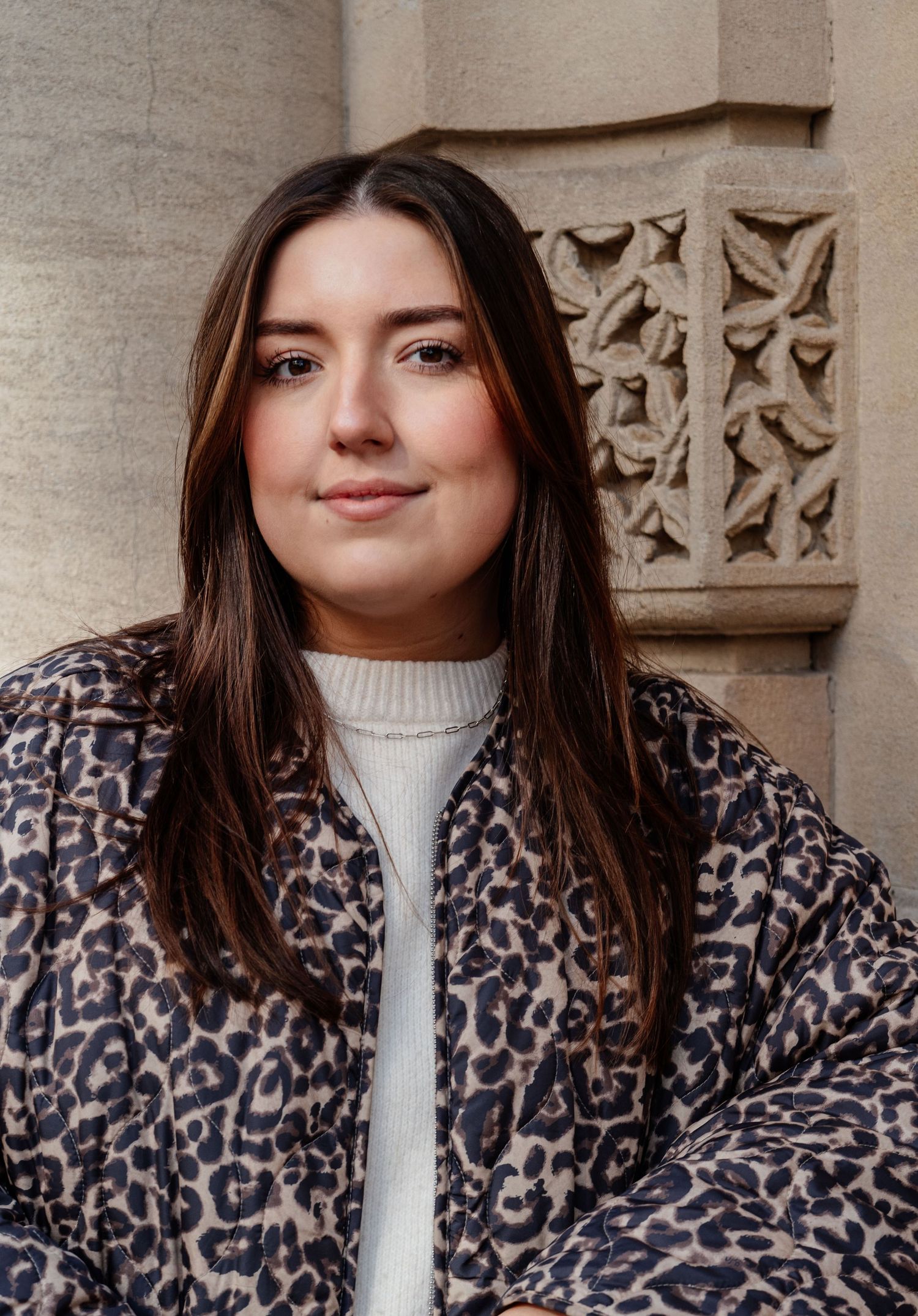 Fashion model wearing a leopard print coat and white turtleneck sweater against a stone wall background.