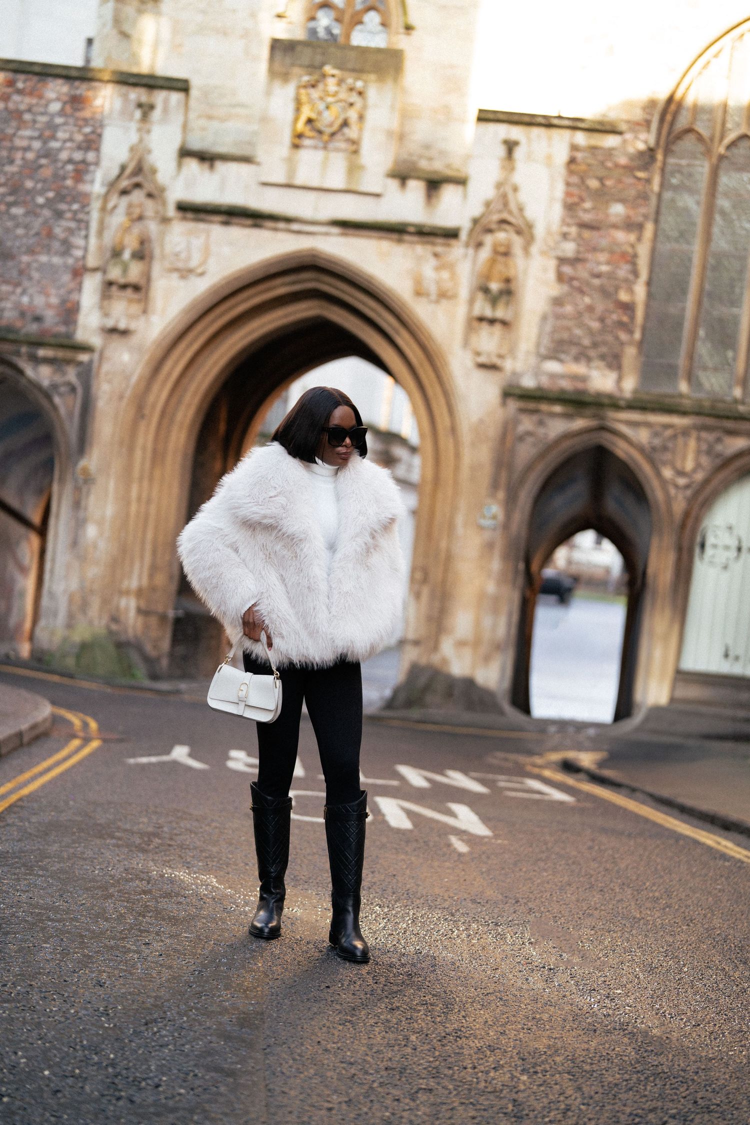 A person wearing a white fur jacket and black pants poses dramatically on a cobblestone street near a historic archway.