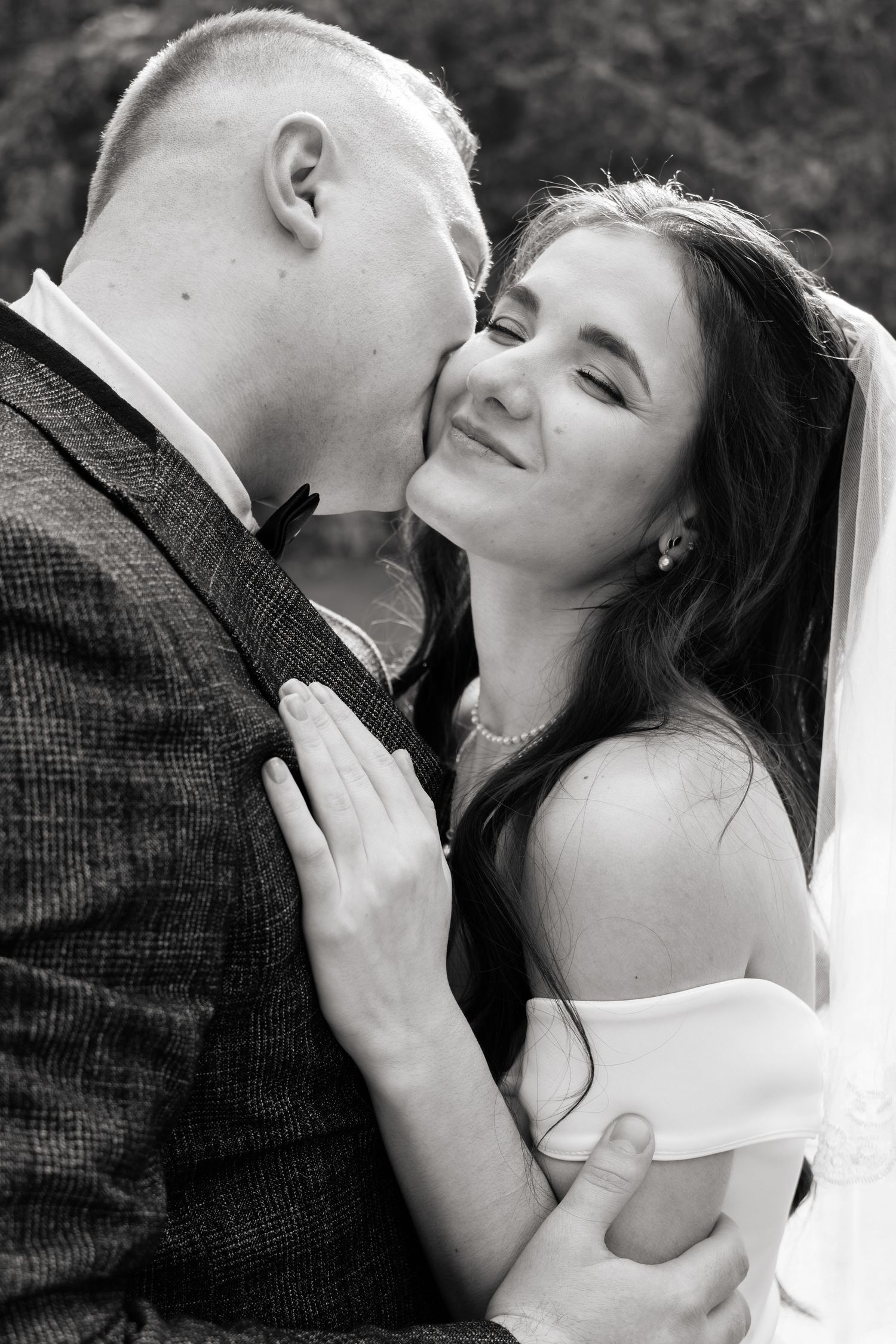 Black and white photo of a groom kissing his smiling bride on the cheek during their wedding day.