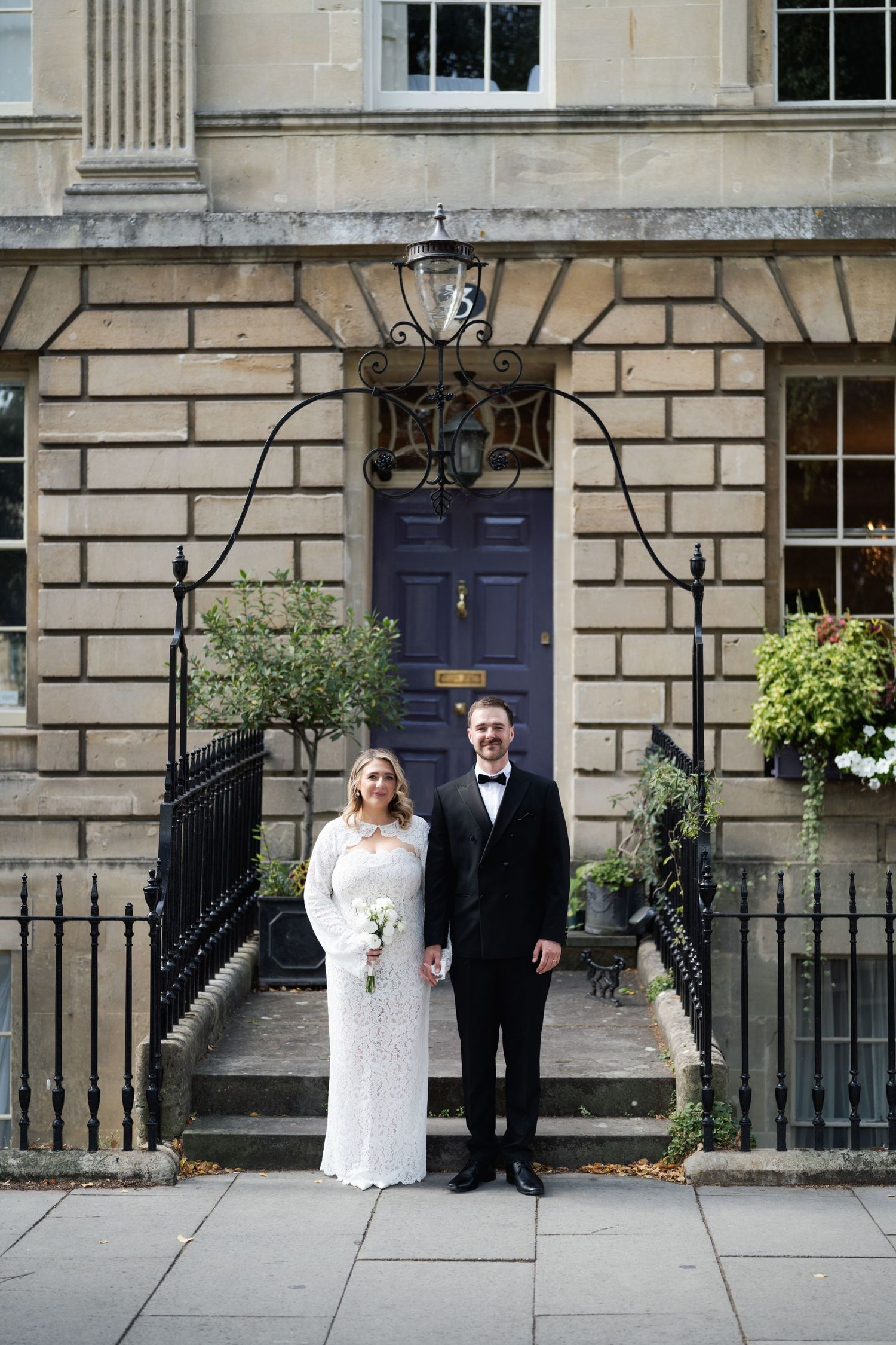 Bride and groom stand before a Georgian stone building with a navy door, she in lace gown, he in black tuxedo.