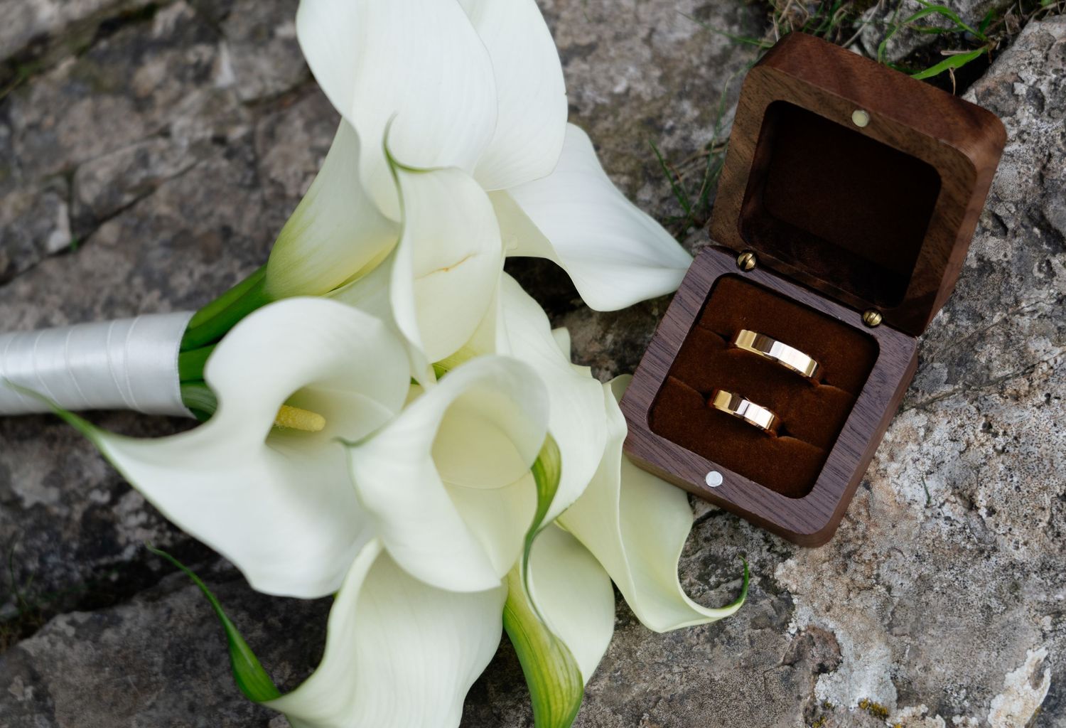 Wedding rings displayed with elegant white calla lilies on stone surface.