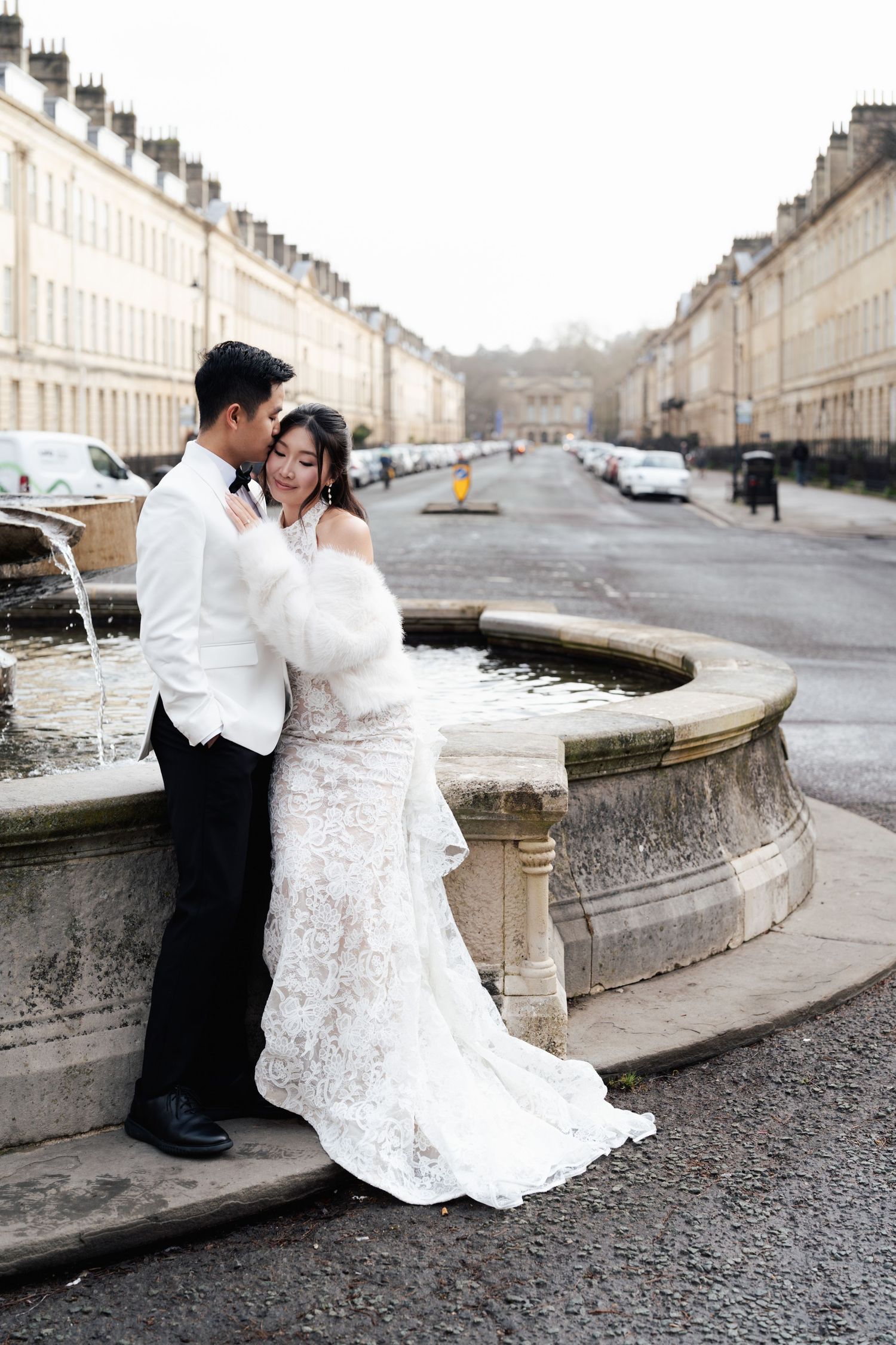 Bride and groom in wedding attire embrace by a stone fountain on a Georgian street in Bath, England.