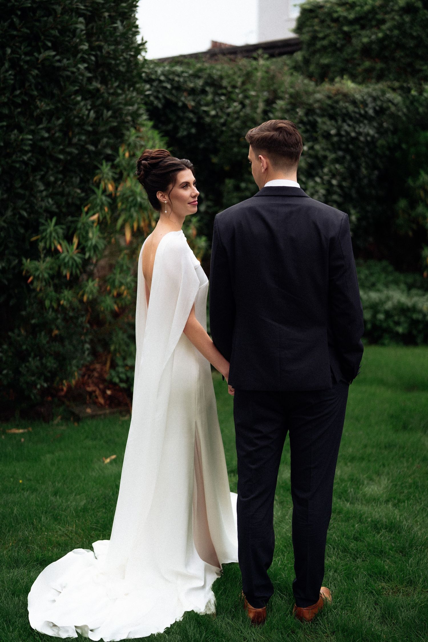 A bride and groom stand together on a green lawn with their backs to the camera, her flowing white veil trailing behind.