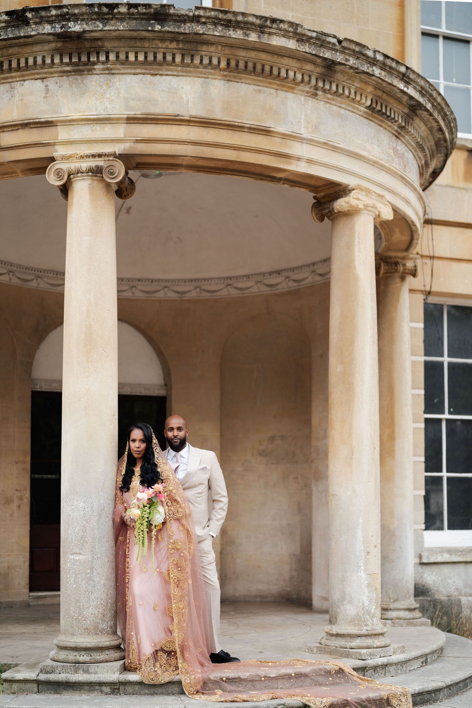 Couple in wedding attire posing under classical stone columns at an elegant historic venue.