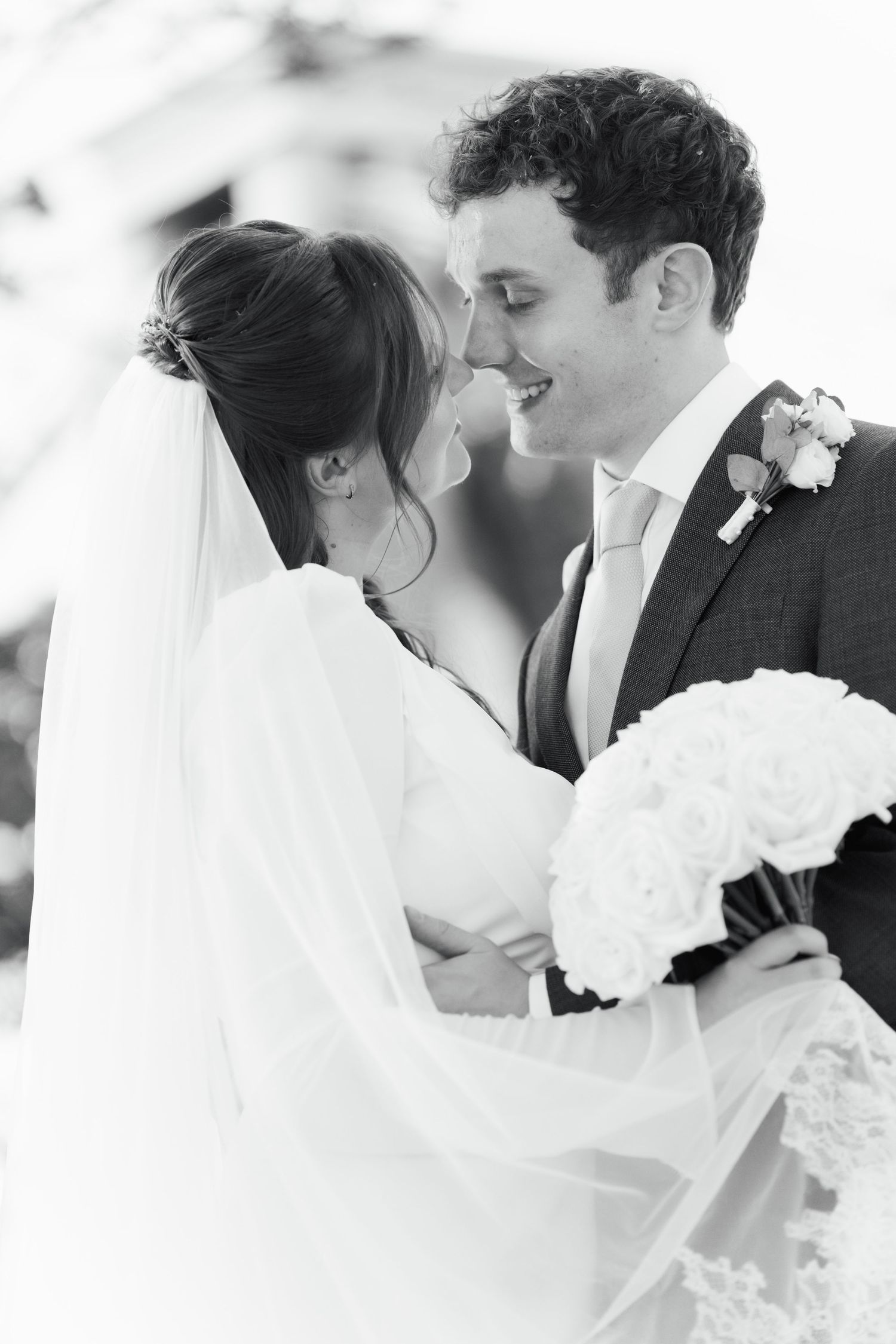 Black and white photo of a smiling groom and bride in veil sharing a romantic moment, holding a white bouquet.