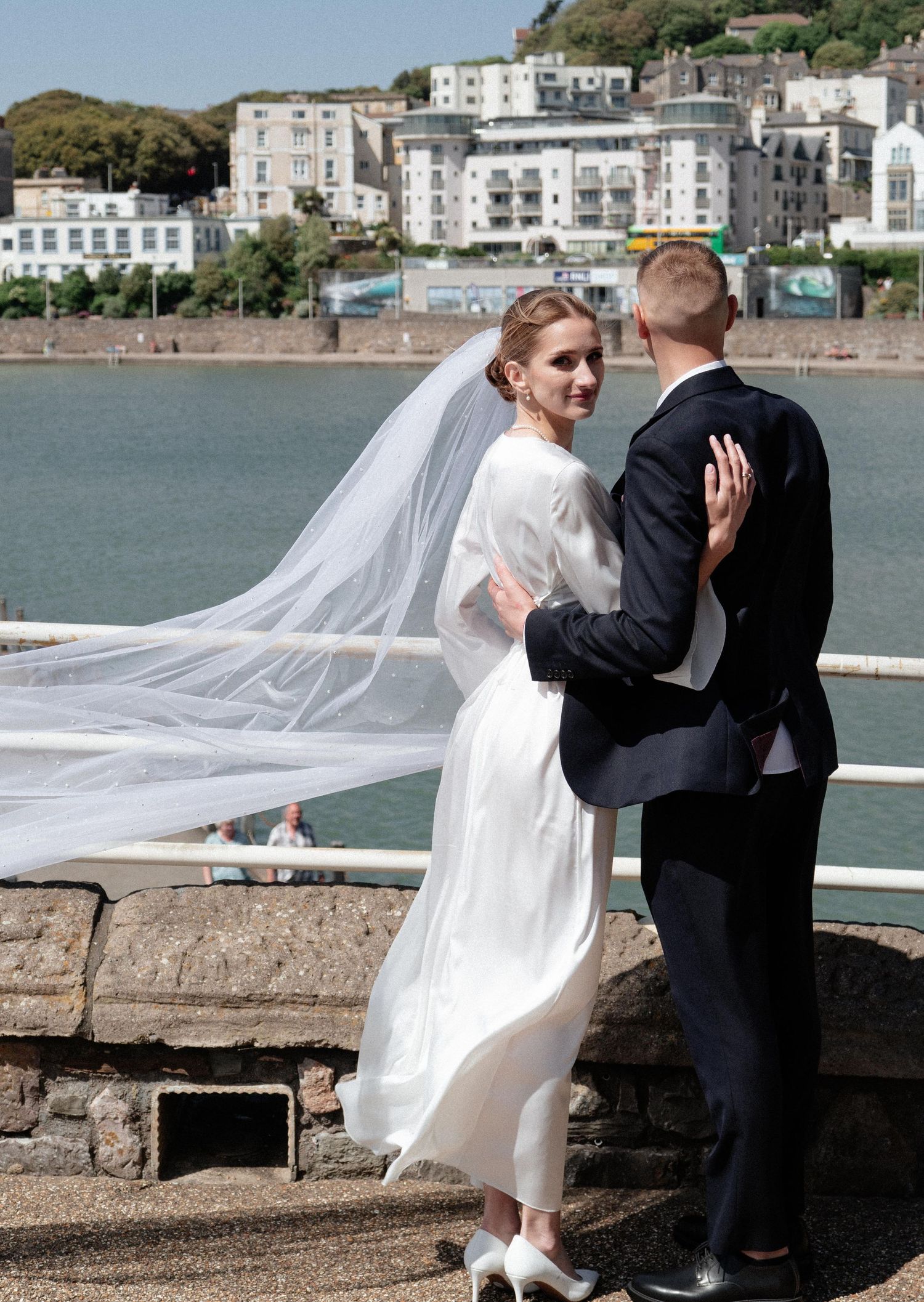 Wedding couple embraces on seaside wall with flowing veil and coastal buildings in background.