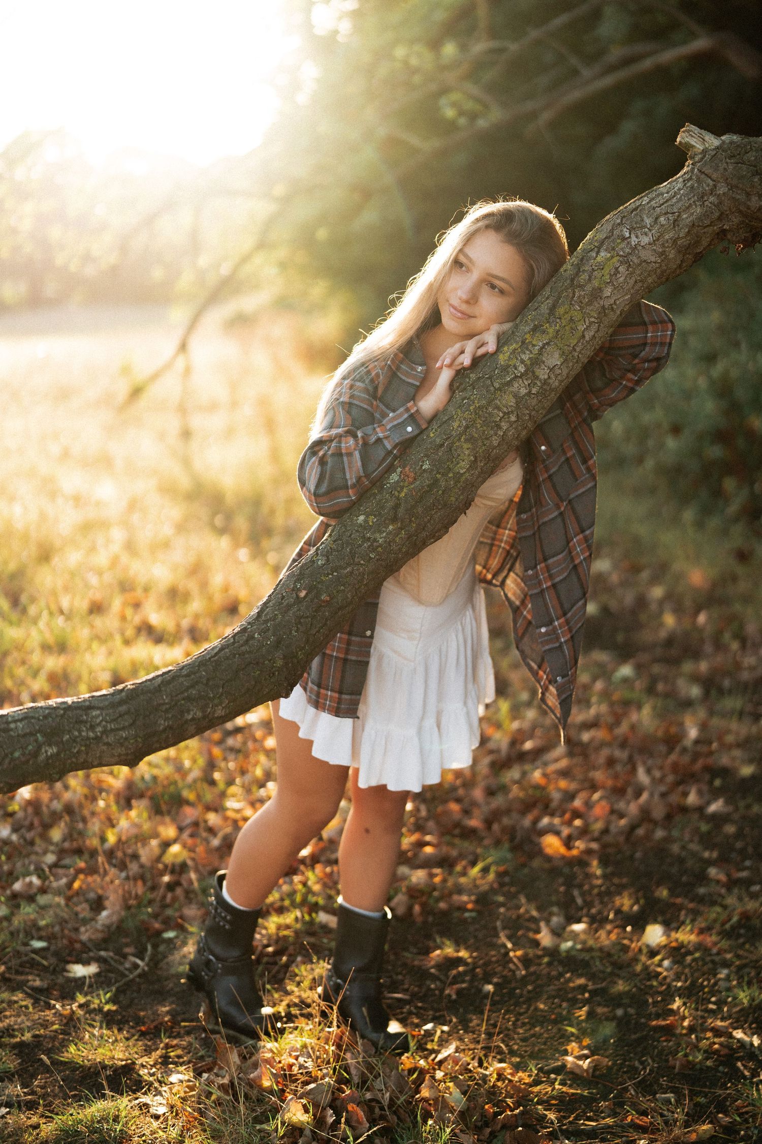 Person in casual attire posing playfully near a tree trunk in autumn sunlight.
