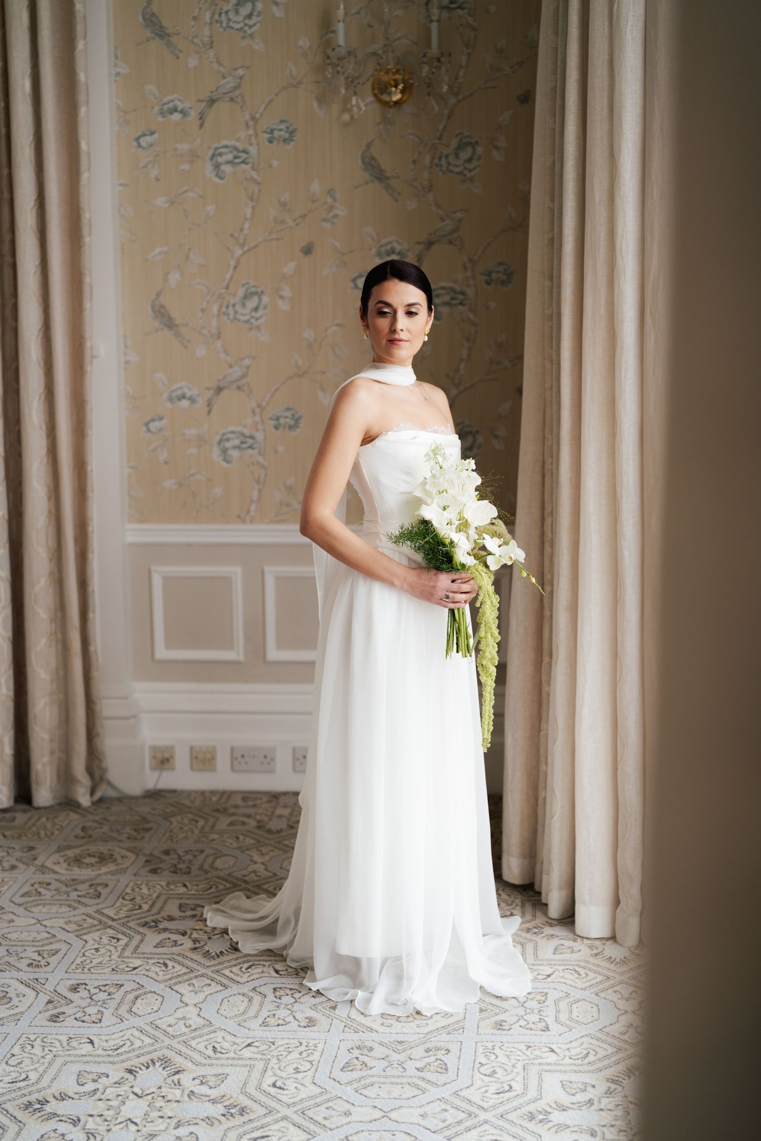 A bride in a flowing white wedding gown holds a classic white bouquet in an elegant room with patterned floors.