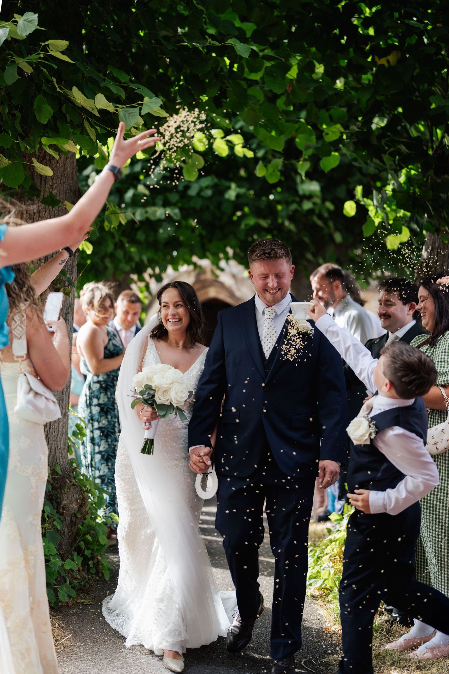 Bride and groom walk through confetti shower surrounded by smiling wedding guests outdoors under green trees.