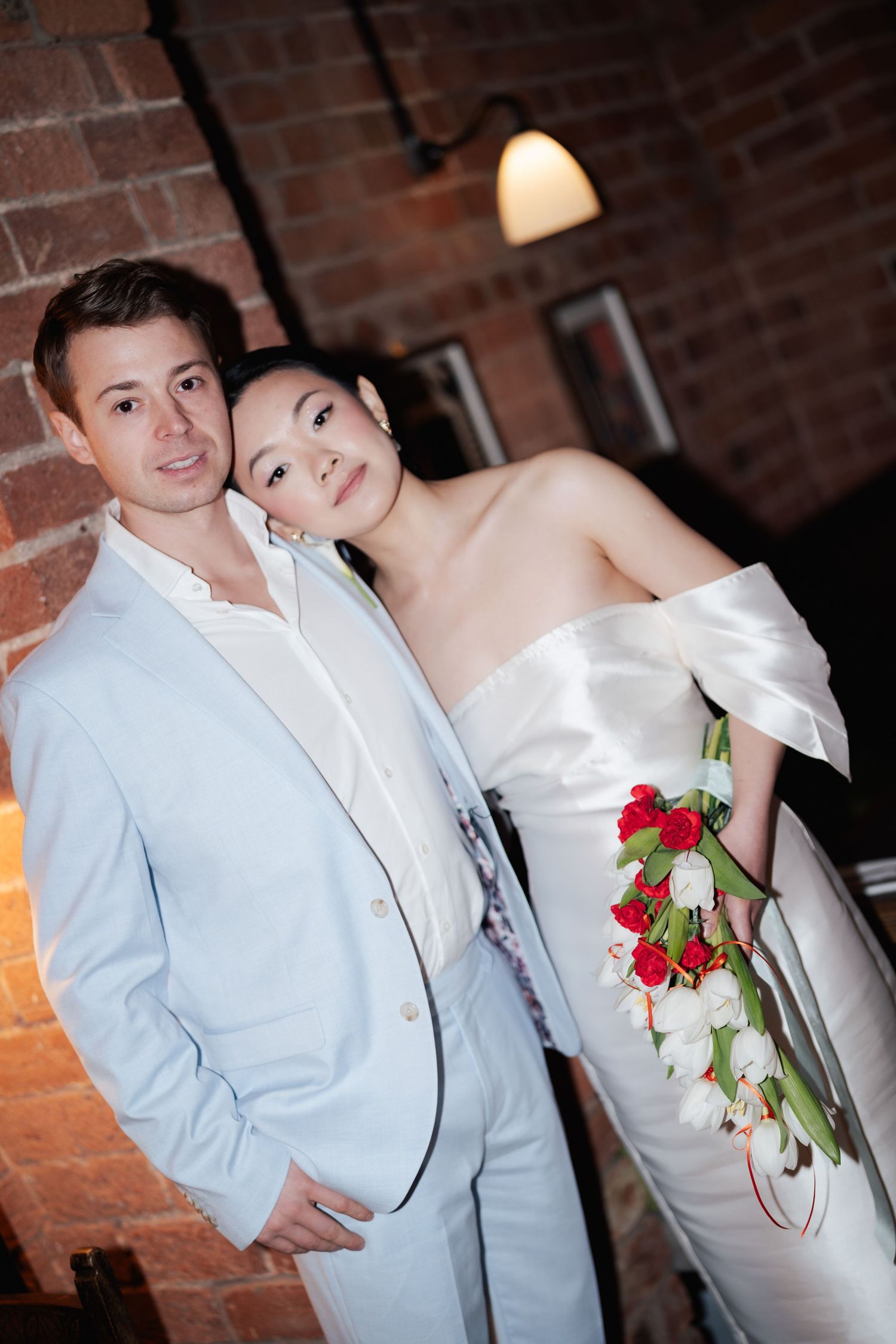 A couple poses for wedding portrait, groom in light blue suit, bride in white off-shoulder gown holding red and white bouquet.