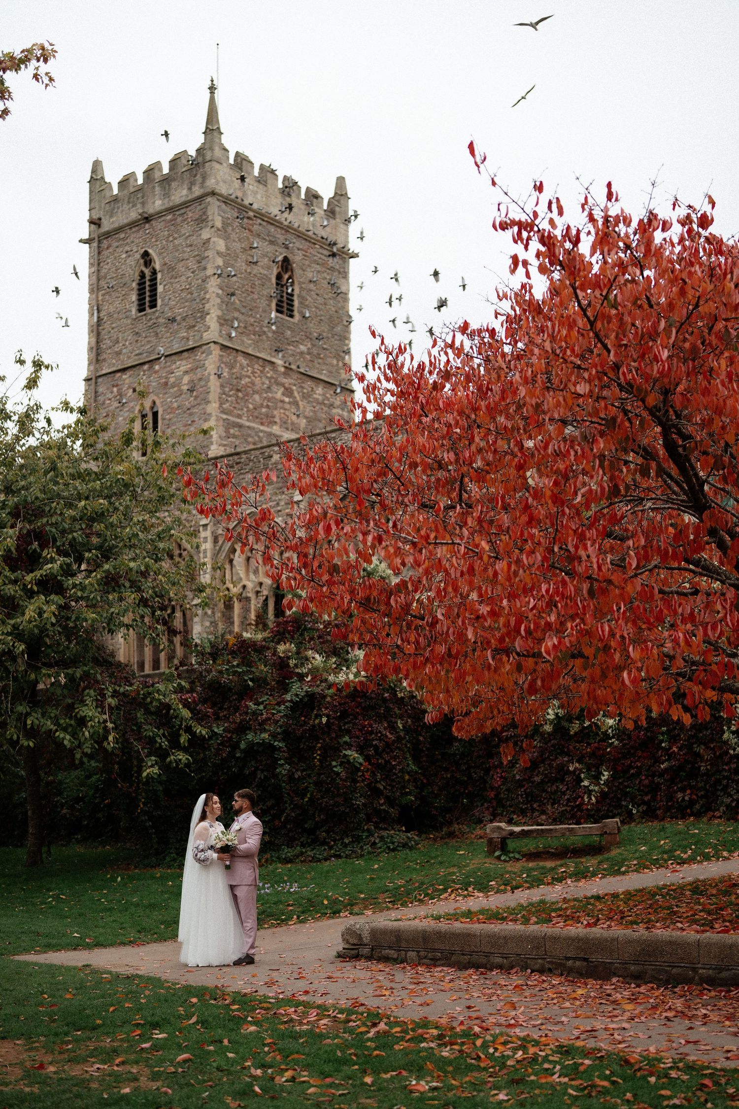 Wedding couple walking near historic stone church with vibrant red autumn trees.