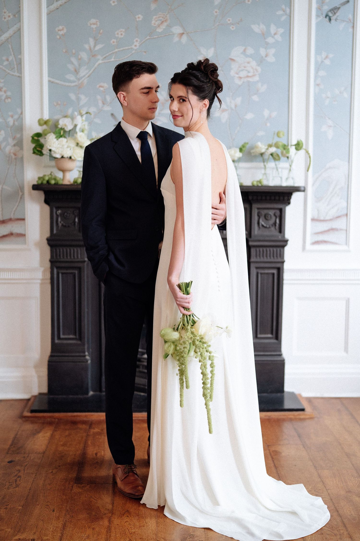 A bride and groom share an intimate moment in an elegant room with blue floral wallpaper and hardwood floors.
