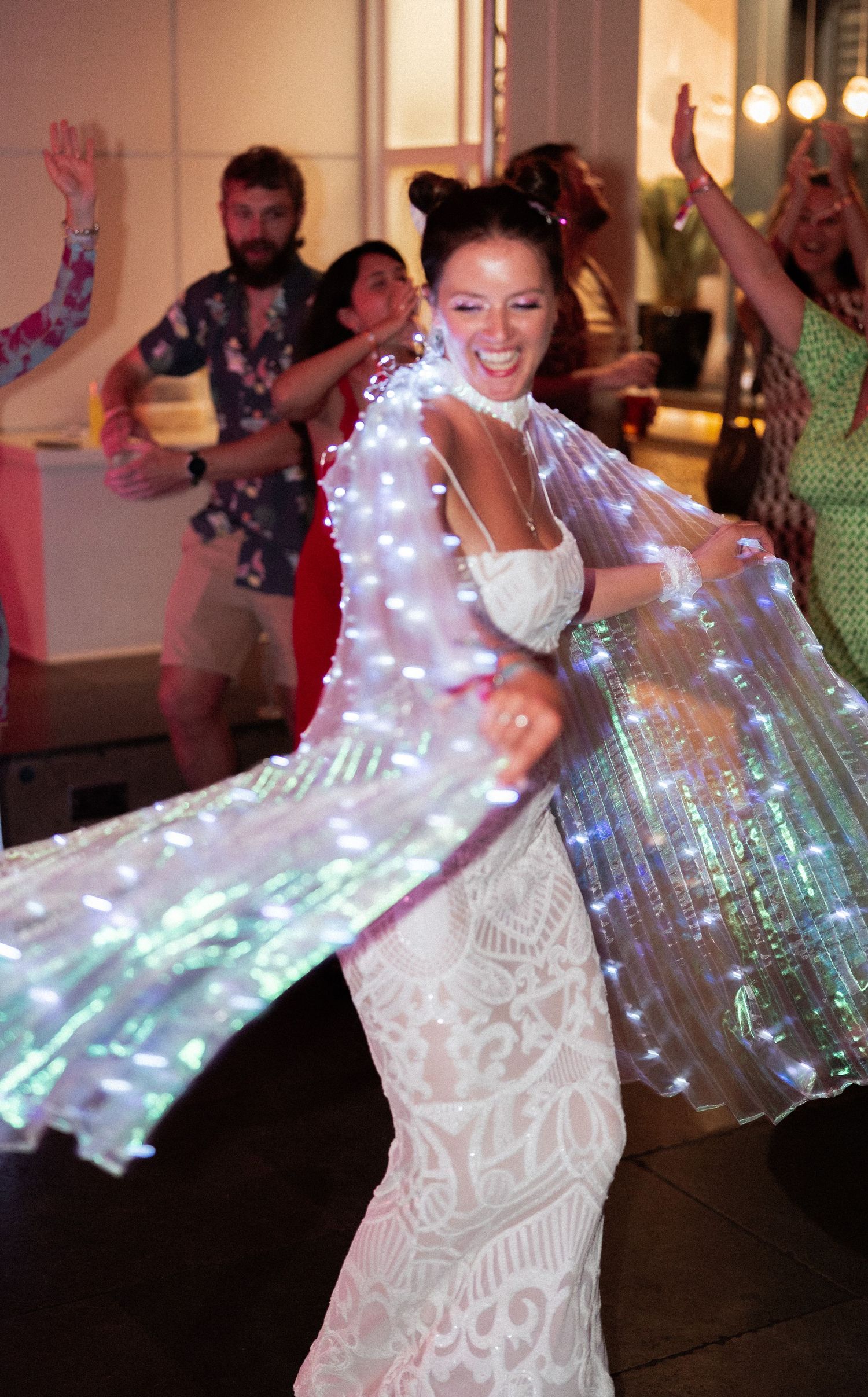 A bride dances joyfully in a white lace gown with a glowing fiber optic cape at a wedding reception.