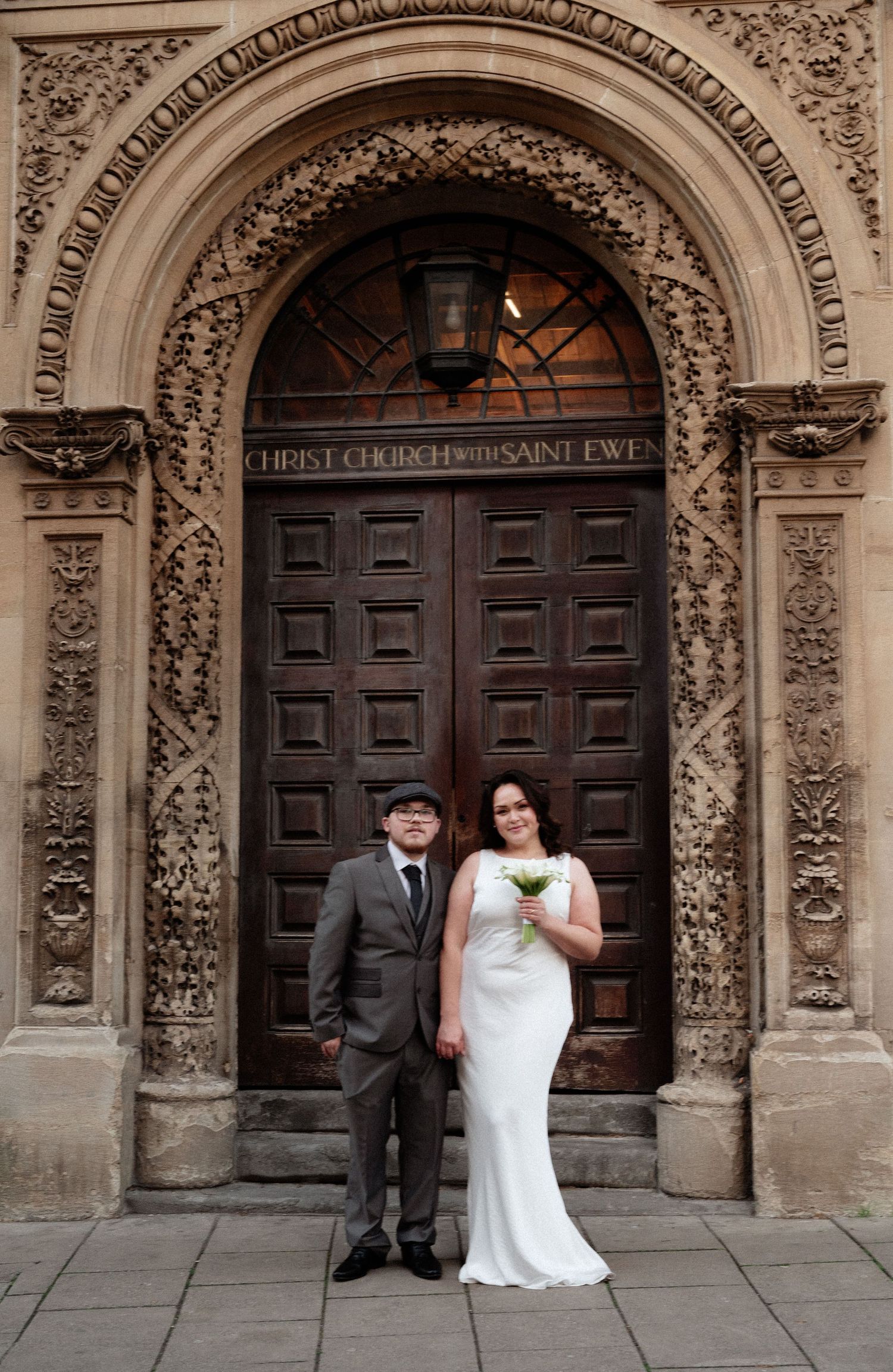A couple poses in front of an ornate stone church entrance with carved archways and wooden doors.