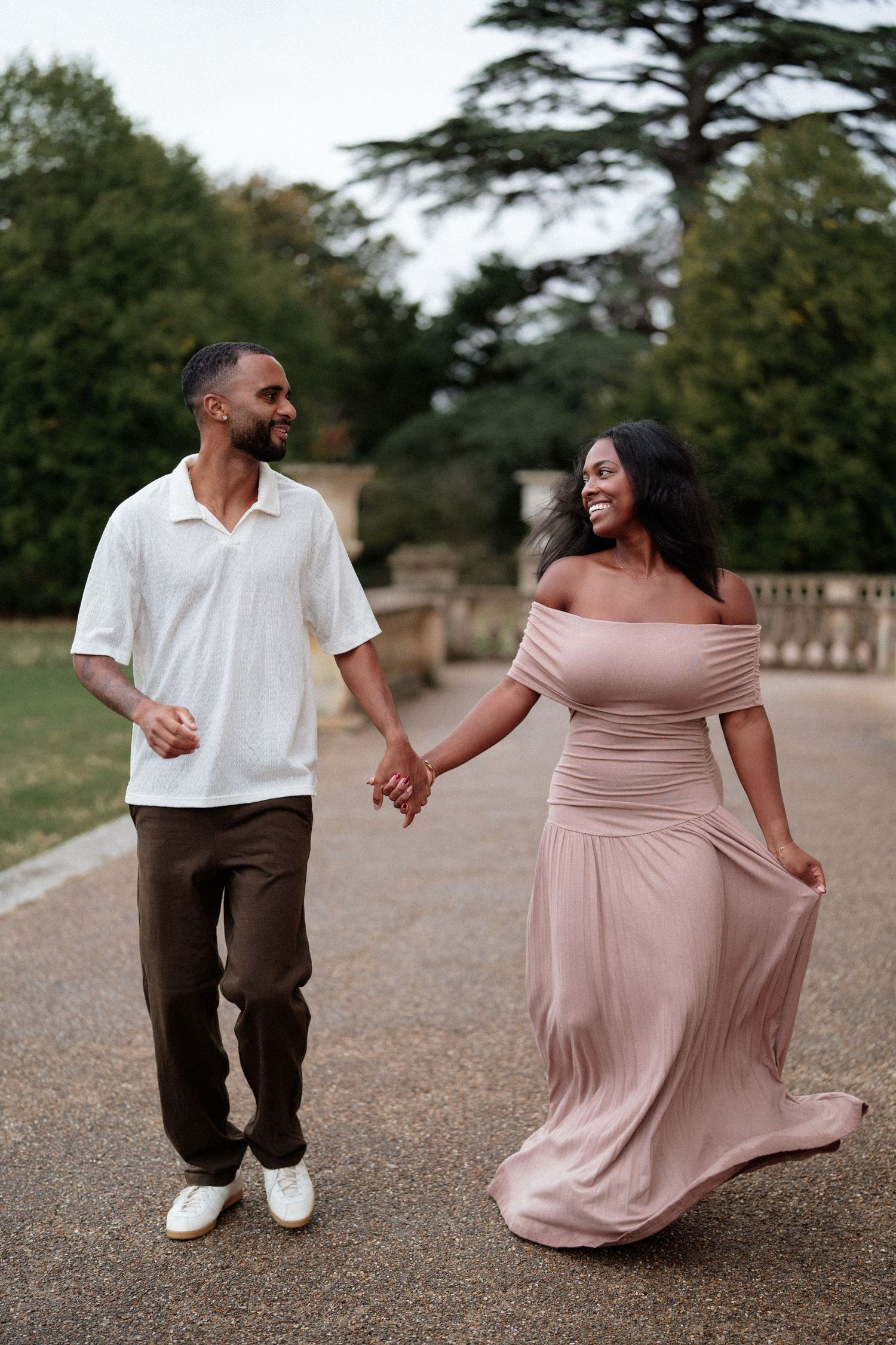 A couple walks hand in hand along a garden path, she wearing a flowing pink dress.
