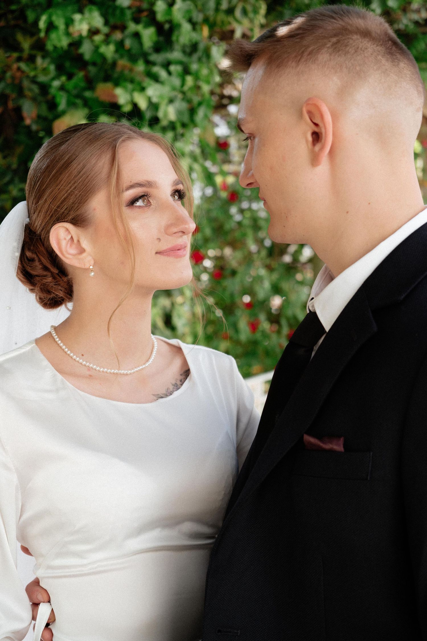 A bride and groom in formal wedding attire gaze lovingly at each other outdoors with green foliage behind them.