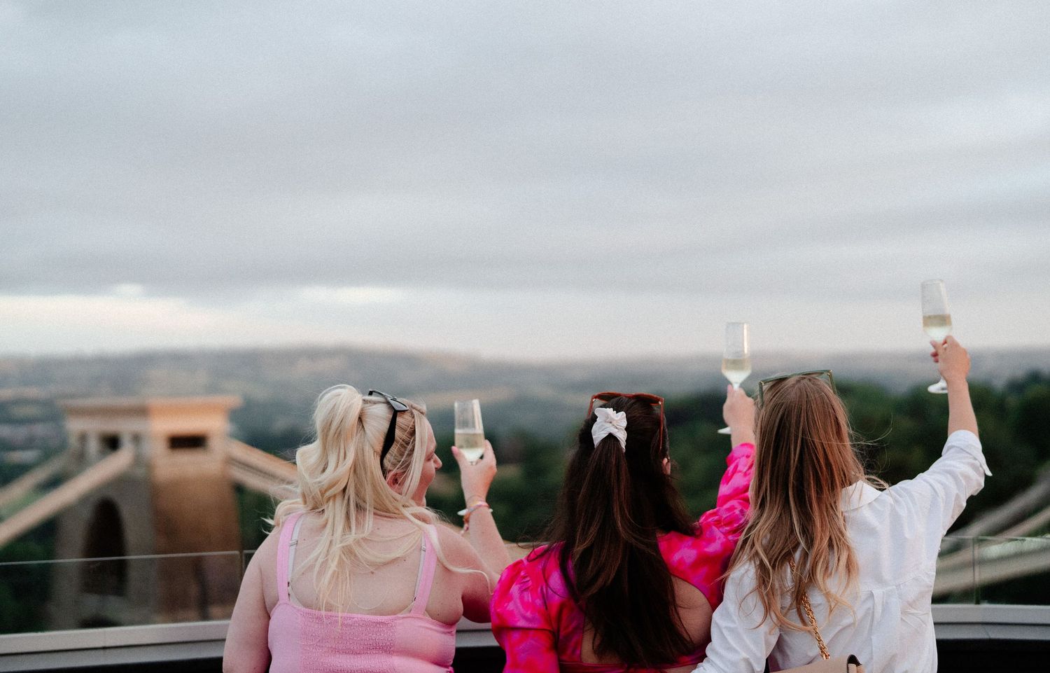 A group of friends in formal attire lean against a railing overlooking a scenic mountain landscape at sunset.
