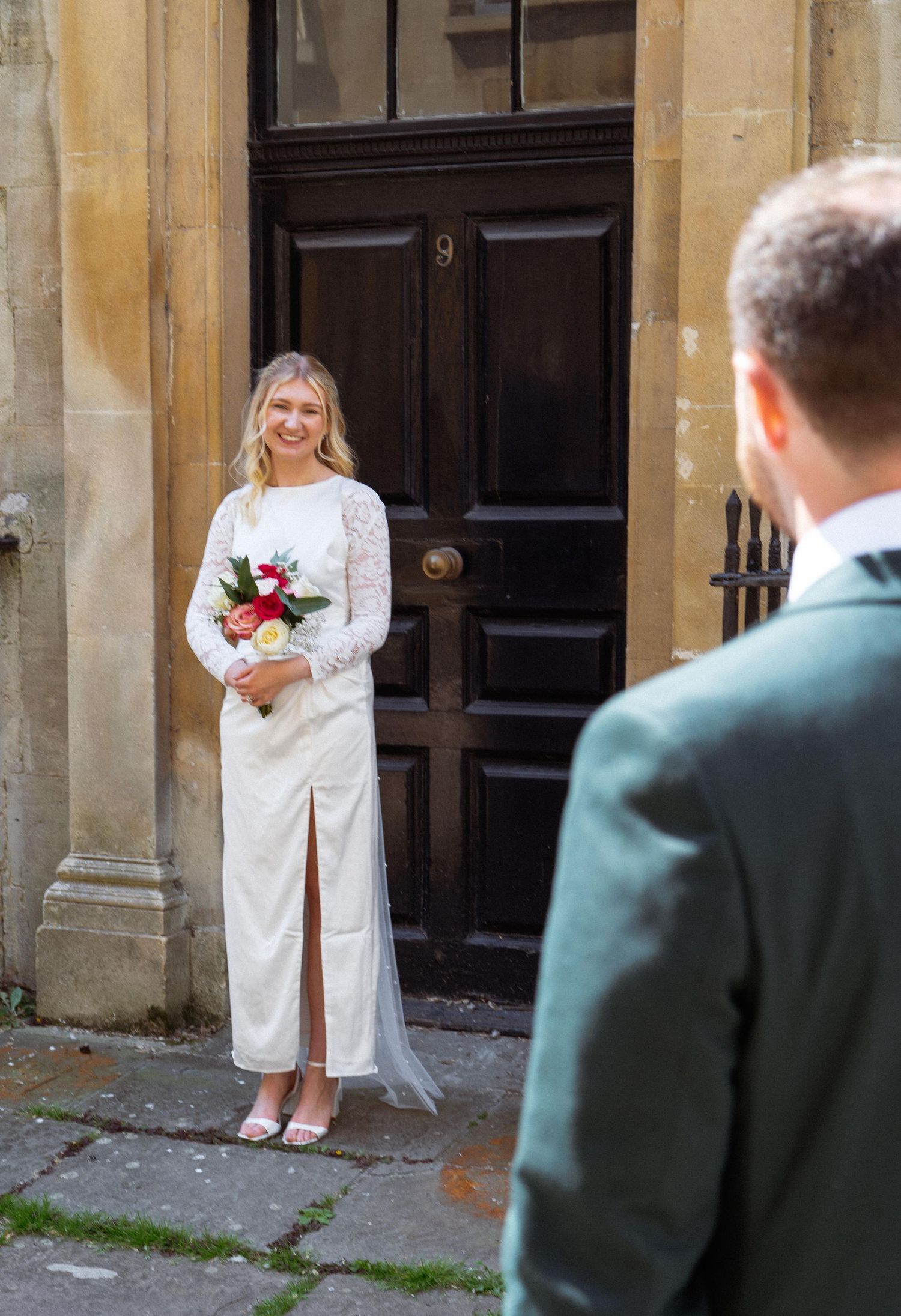 A bride in a white lace dress with a thigh slit holds a bouquet while standing in front of a dark wooden door.