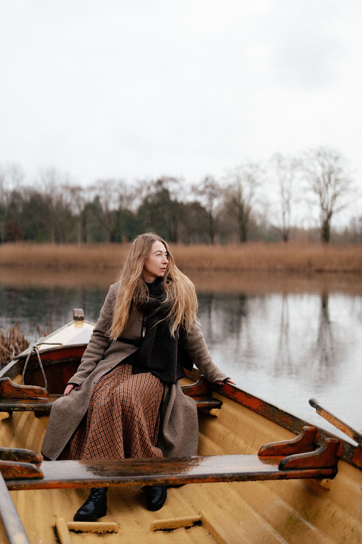 A person in a brown coat sits in a wooden rowboat on a calm lake surrounded by bare trees in winter.