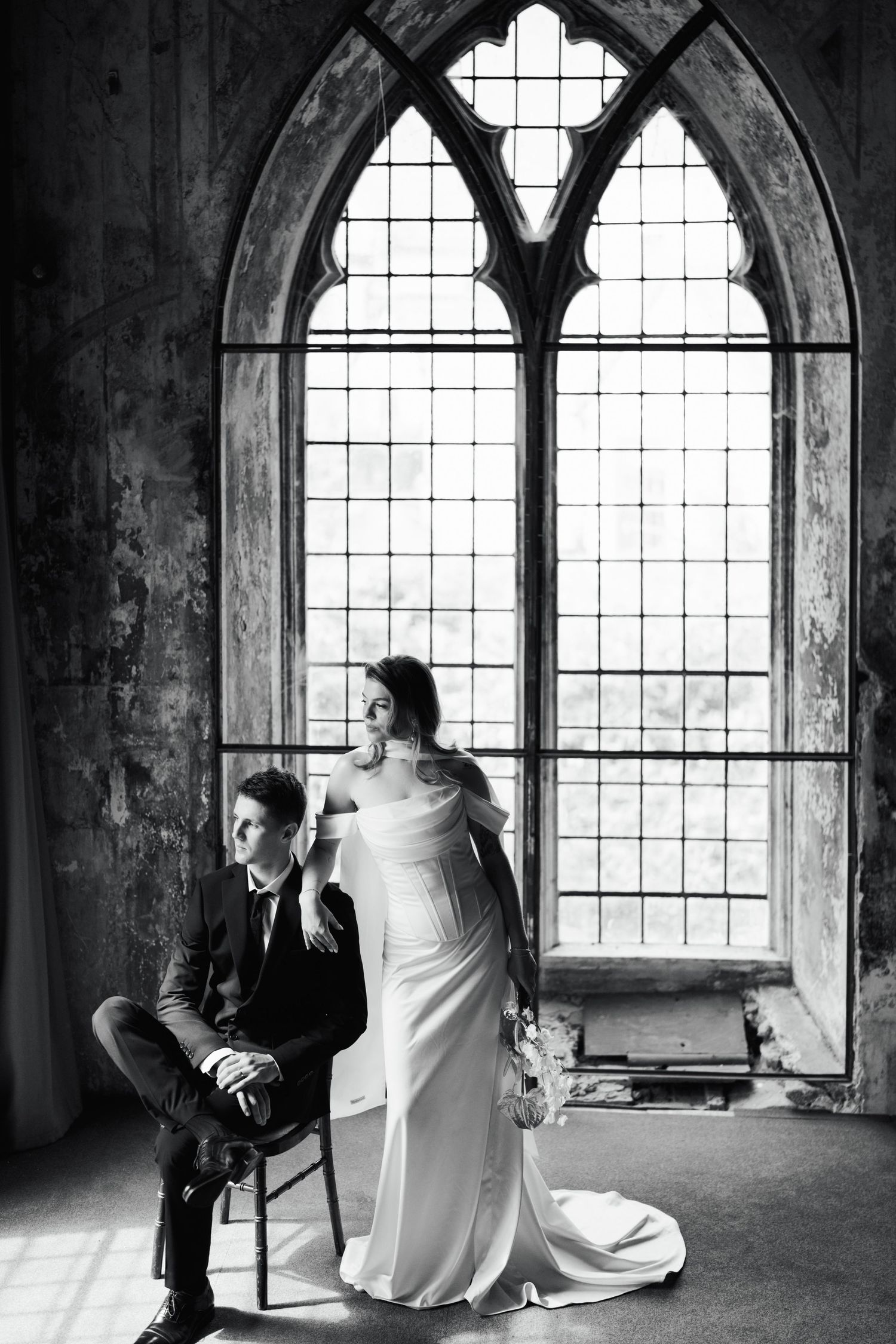 Black and white wedding photo of bride in elegant gown and groom in suit posing by gothic arched window in historic venue.