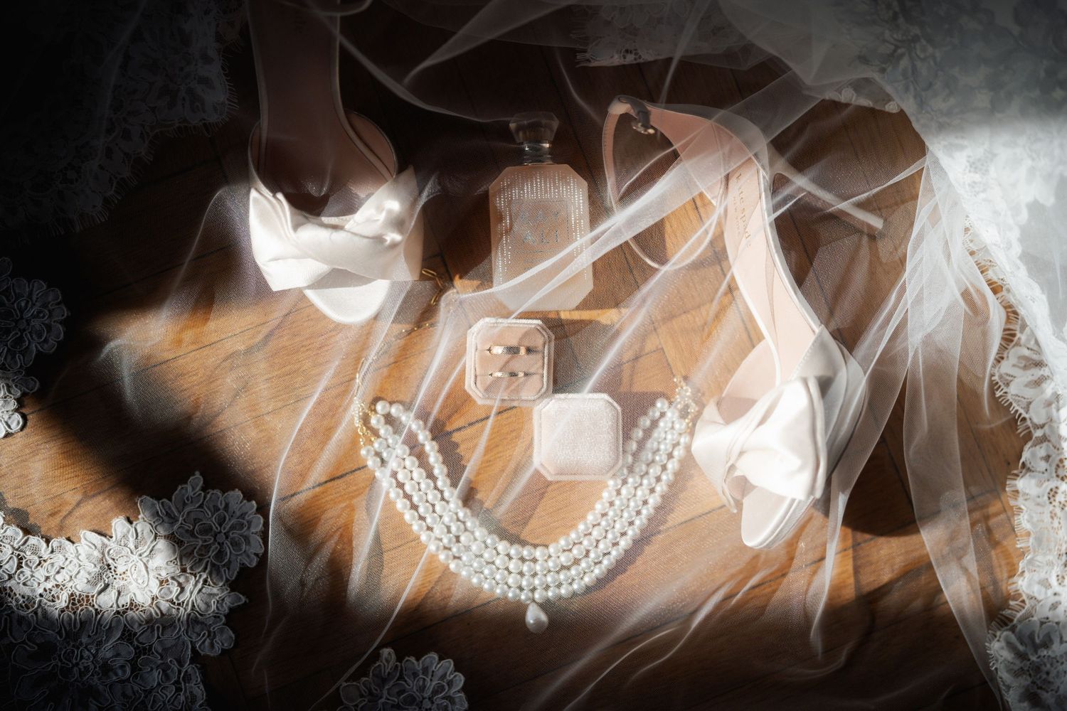 Bridal accessories flat lay with pearl necklace, heels, veil, perfume bottle, and jewelry box on wooden surface.